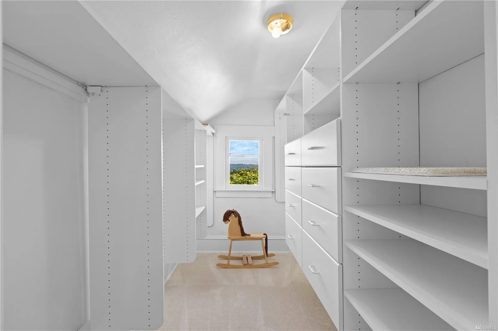 Empty walk-in closet with white shelves and drawers, a rocking horse, a window showing a green landscape, beige carpet, and a ceiling light fixture.