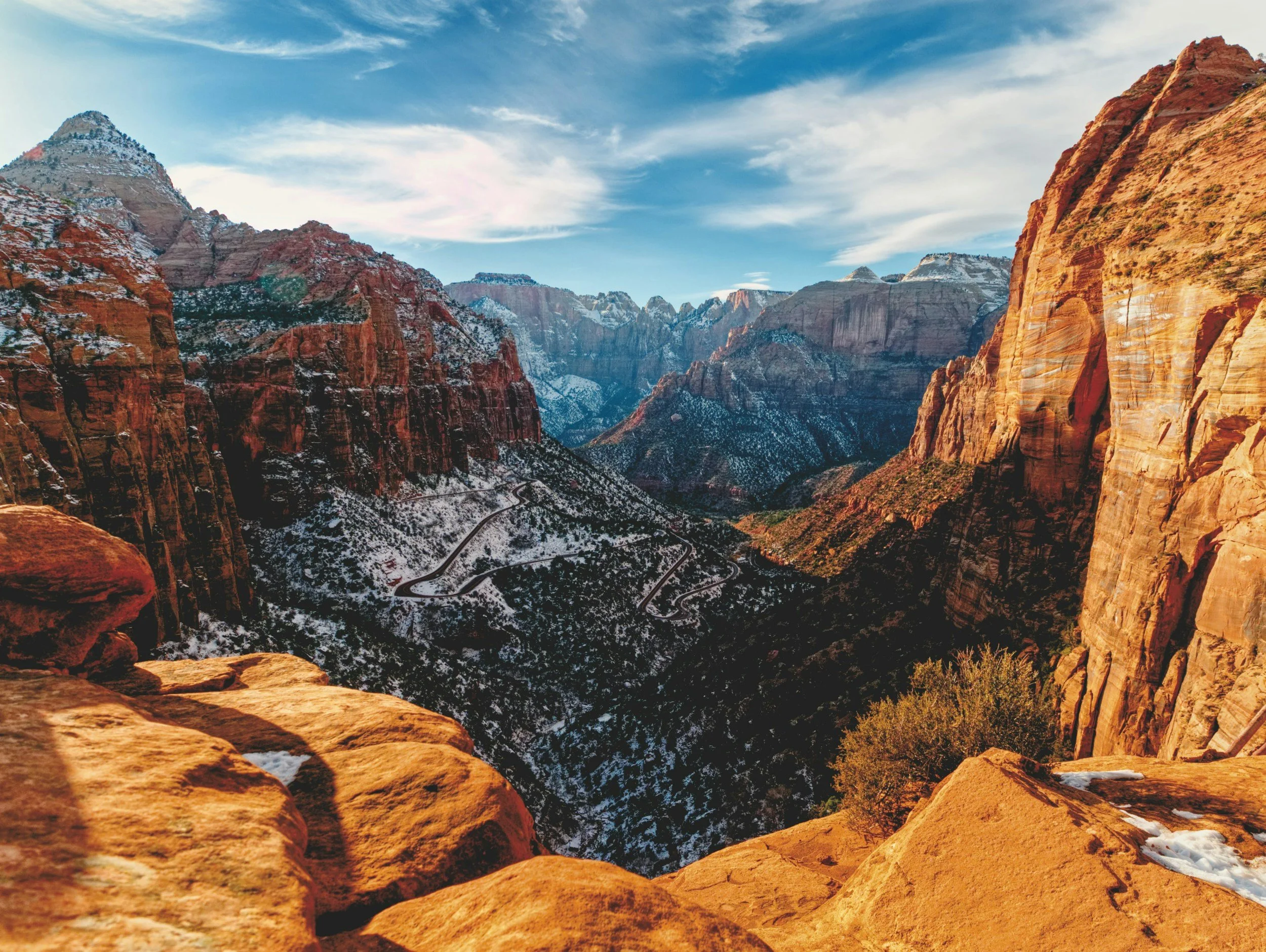 View of Grand Canyon with colorful red and brown cliffs, snowy patches on the ground, winding roads, and a partly cloudy blue sky.
