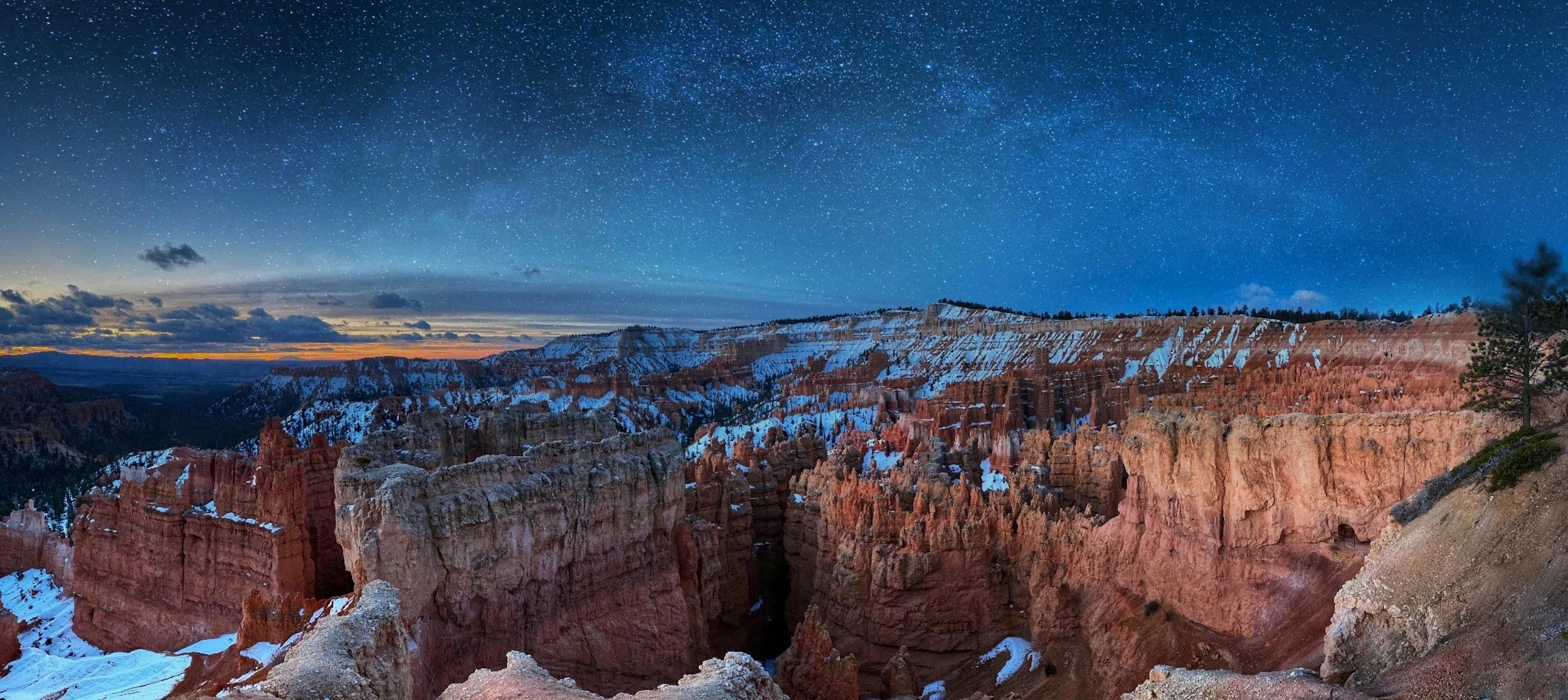 Nighttime view of Bryce Canyon with snow-capped orange and pink hoodoos and a starry sky overhead.