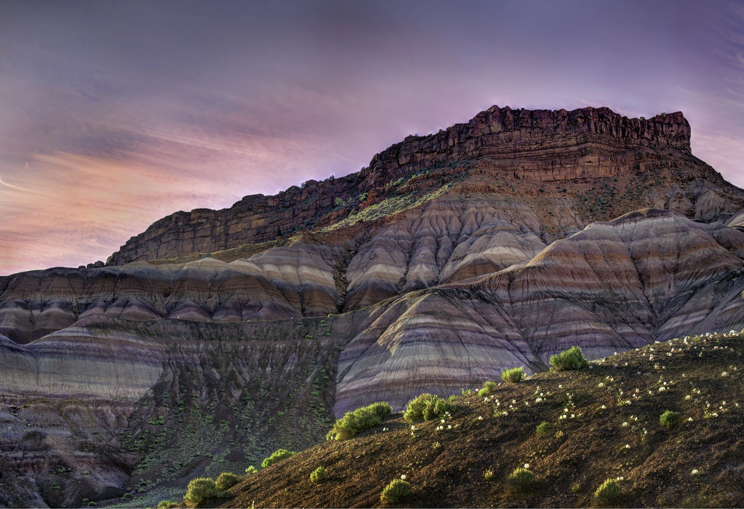 Colorful layered rocks and hills with sparse green vegetation under a pink and purple sky during sunset