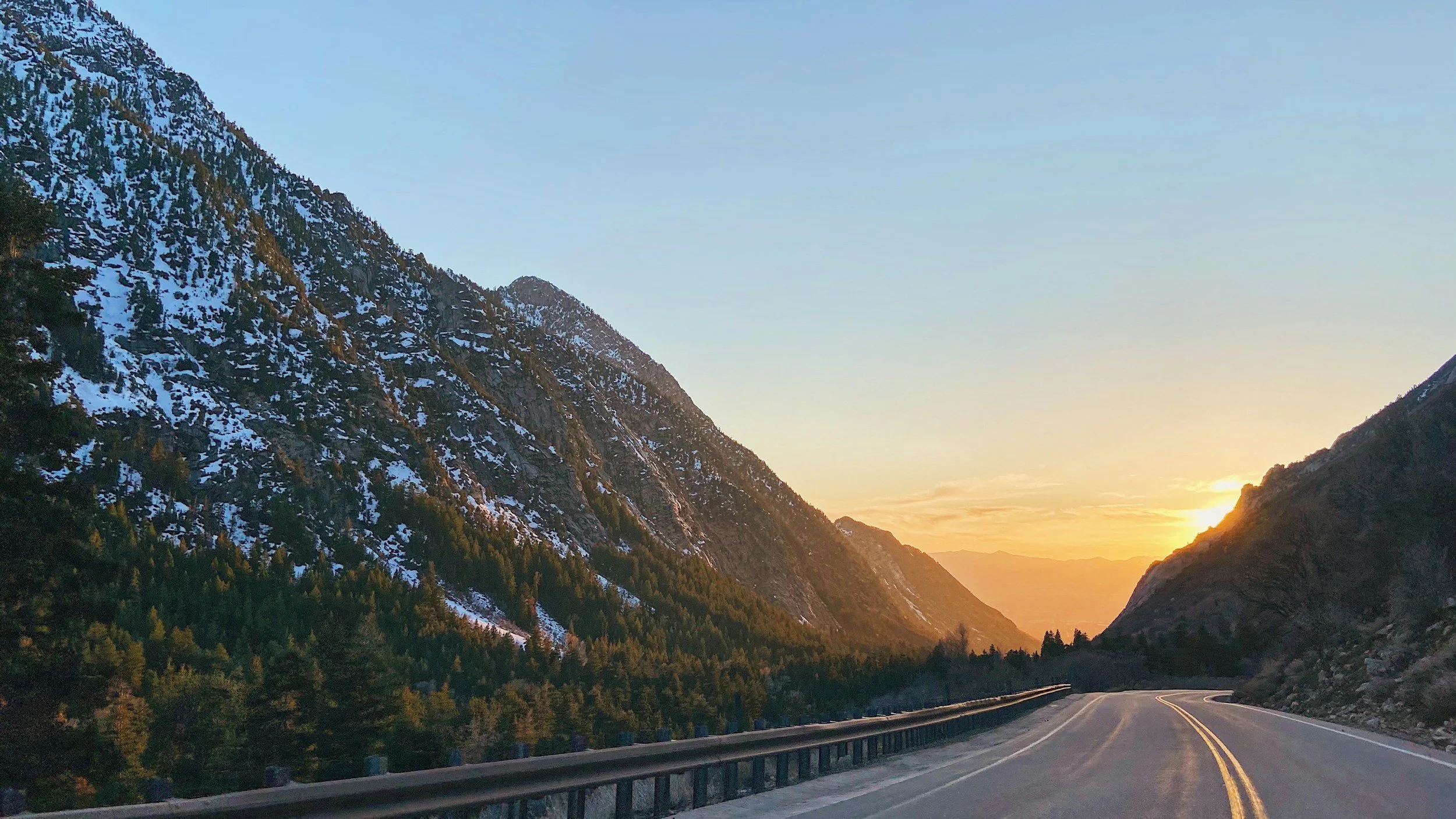 A winding mountain road at sunset with snow-covered mountains and pine trees on either side.