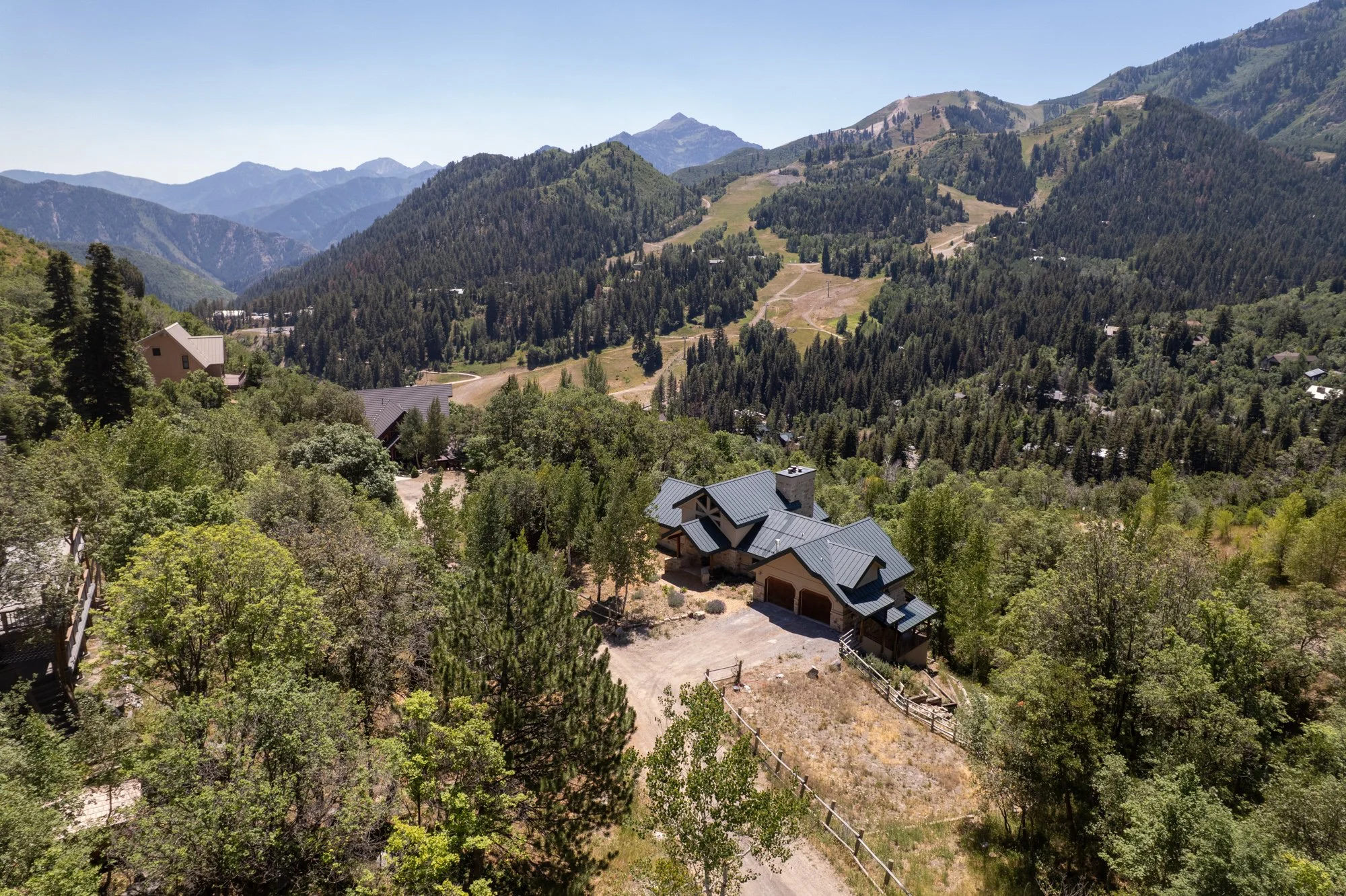 Aerial view of a house with a metal roof in a mountainous forested area, surrounded by trees and other houses, with mountain peaks in the background.