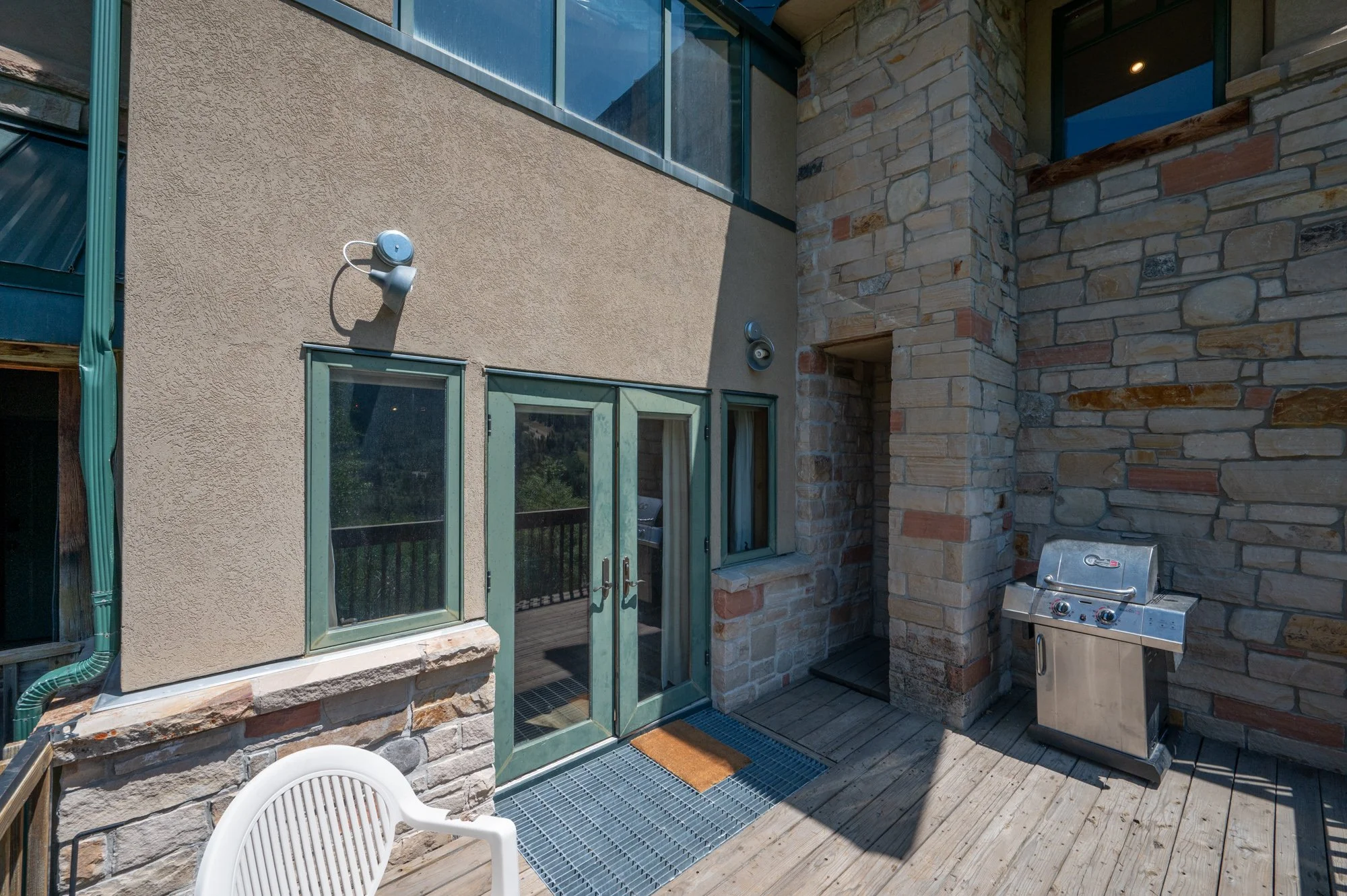 An outdoor patio area with a stainless steel grill, a plastic chair, a glass door leading inside, stone and stucco walls, and a wooden decking surface.