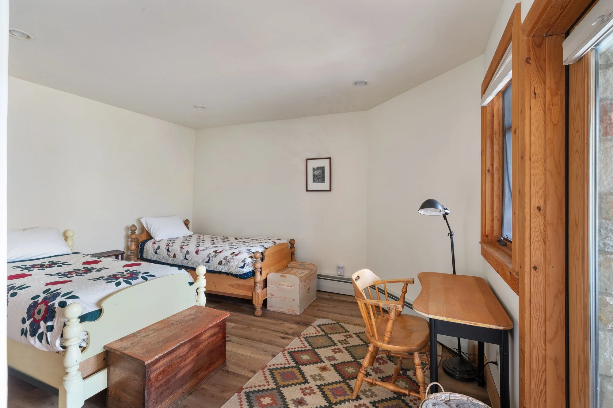A bedroom with two beds, a wooden chest at the foot of one bed, a small wooden desk with a chair, a black adjustable desk lamp, a framed picture on the wall, a window with wooden trim and white blinds, a patterned area rug, and wooden flooring.