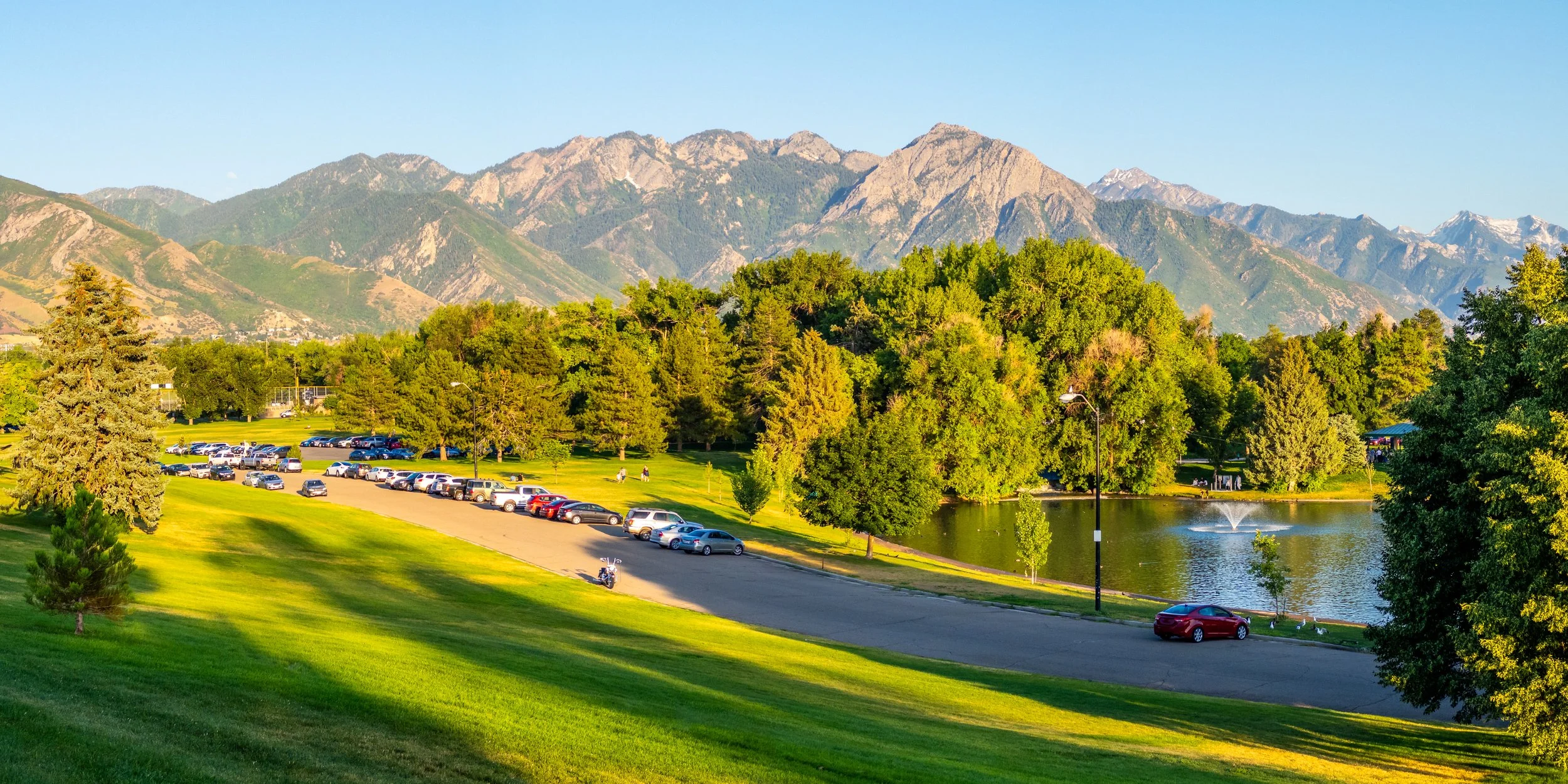 A scenic park with a pond, surrounded by lush trees, with a parking lot filled with cars and a mountain range in the background on a sunny day.