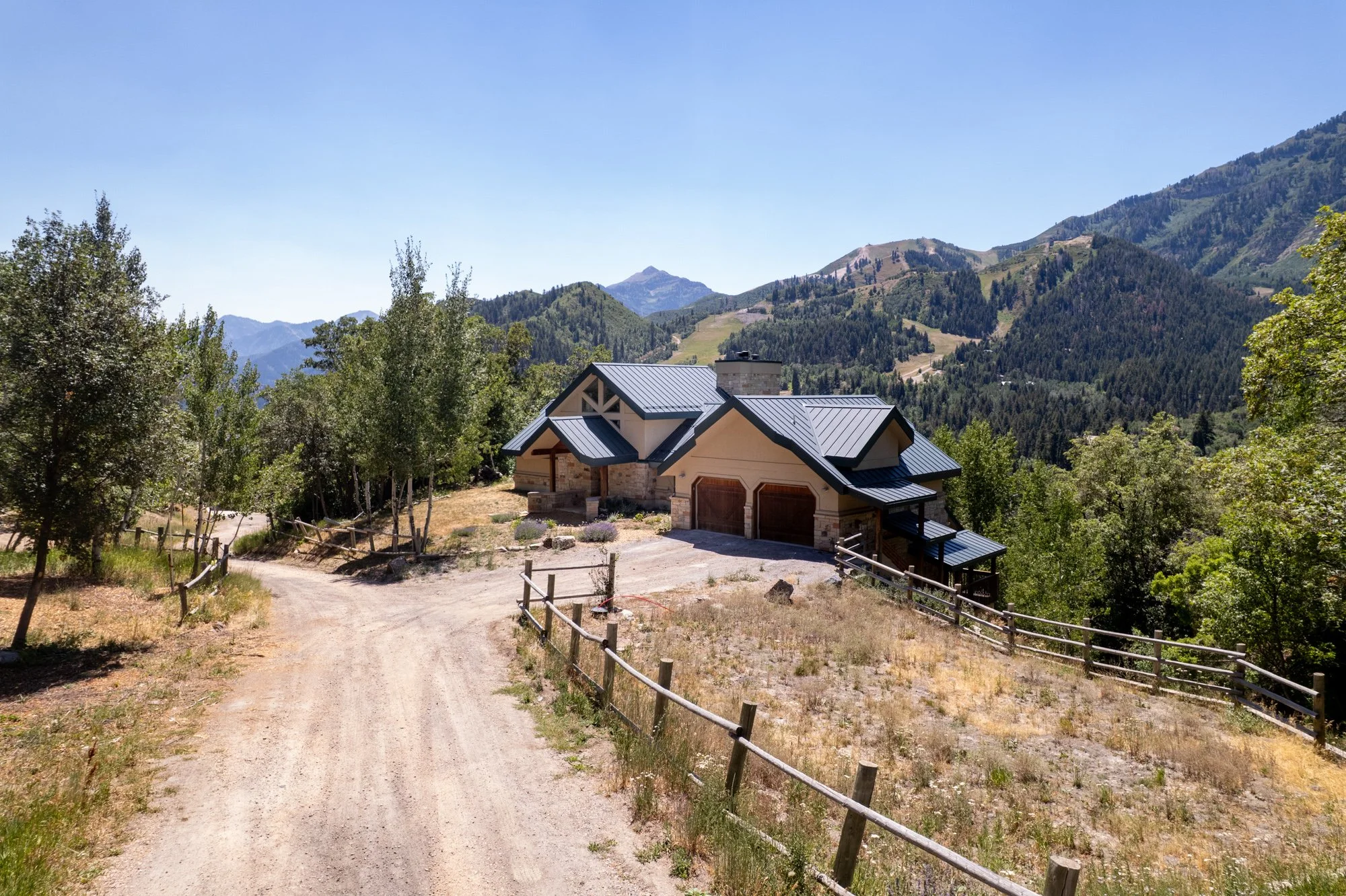 A house with a dark metal roof surrounded by trees and mountains in the background, with a dirt driveway leading up to it.