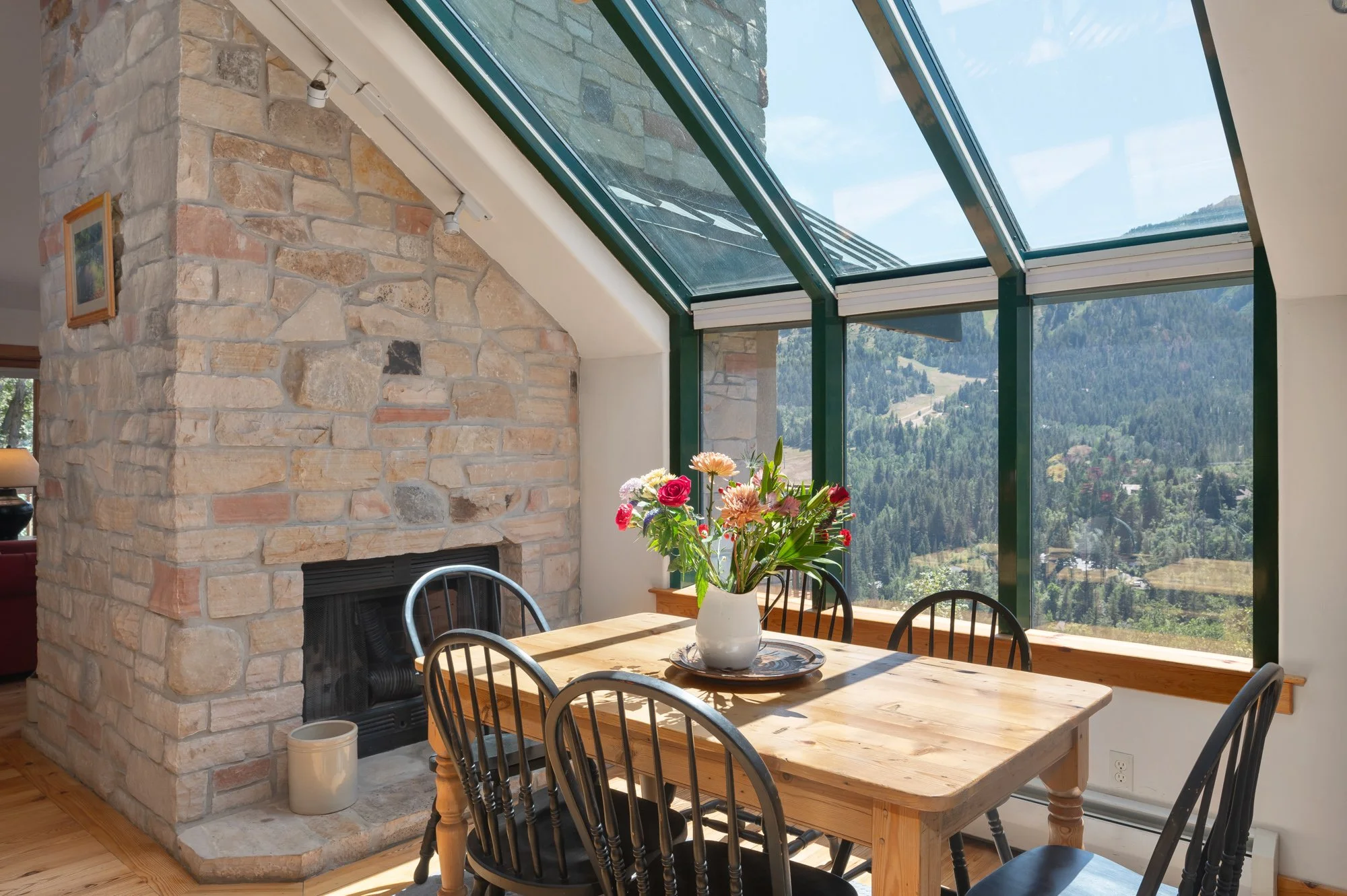 Dining area with a wooden table and six black chairs in front of a large window and a stone fireplace. A white vase with colorful flowers sits on the table. The large window provides a view of a mountainous landscape.