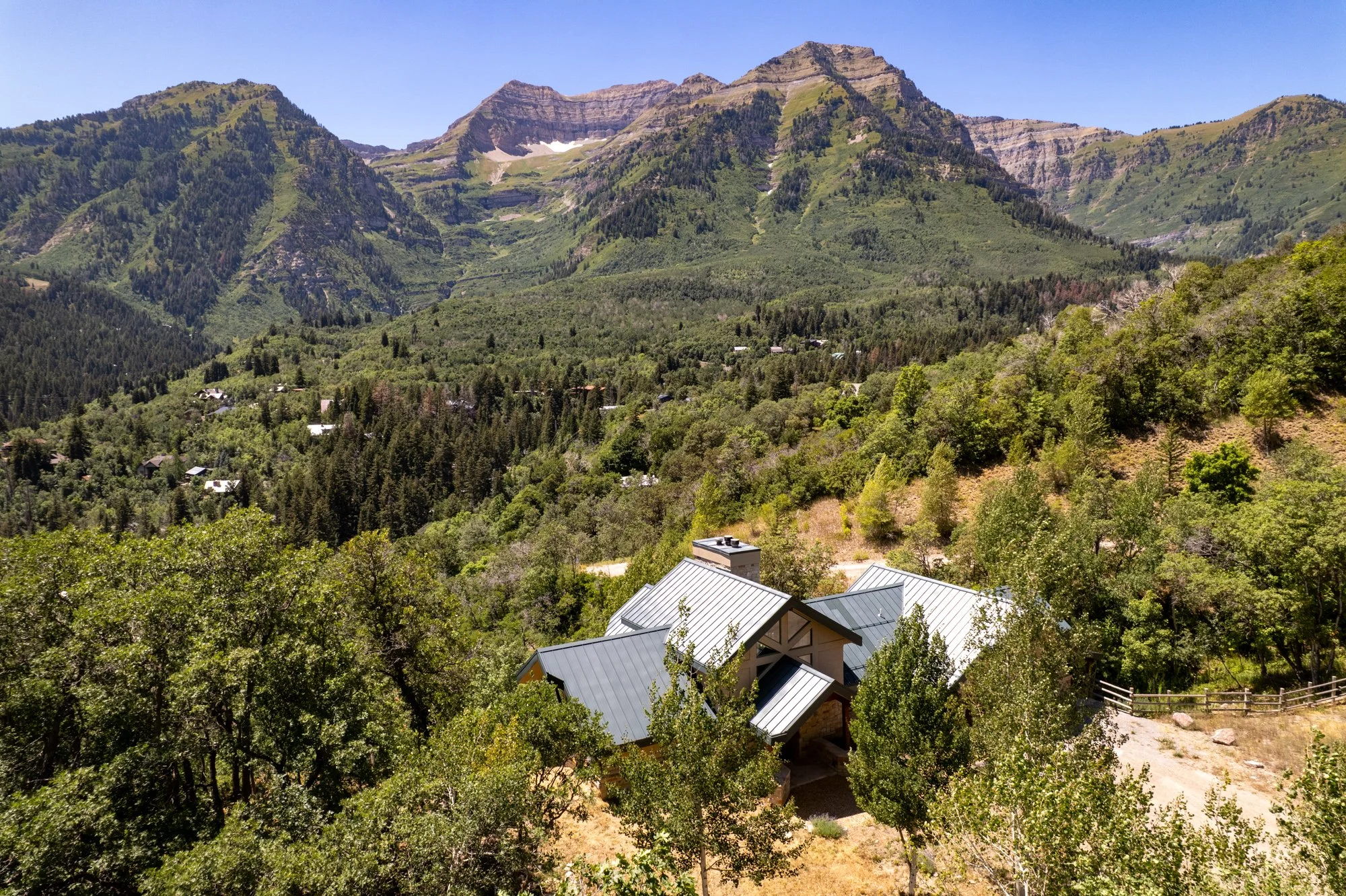 A house with metal roofing nestled among green trees in a mountain valley, surrounded by forested hills and rugged mountains in the background.