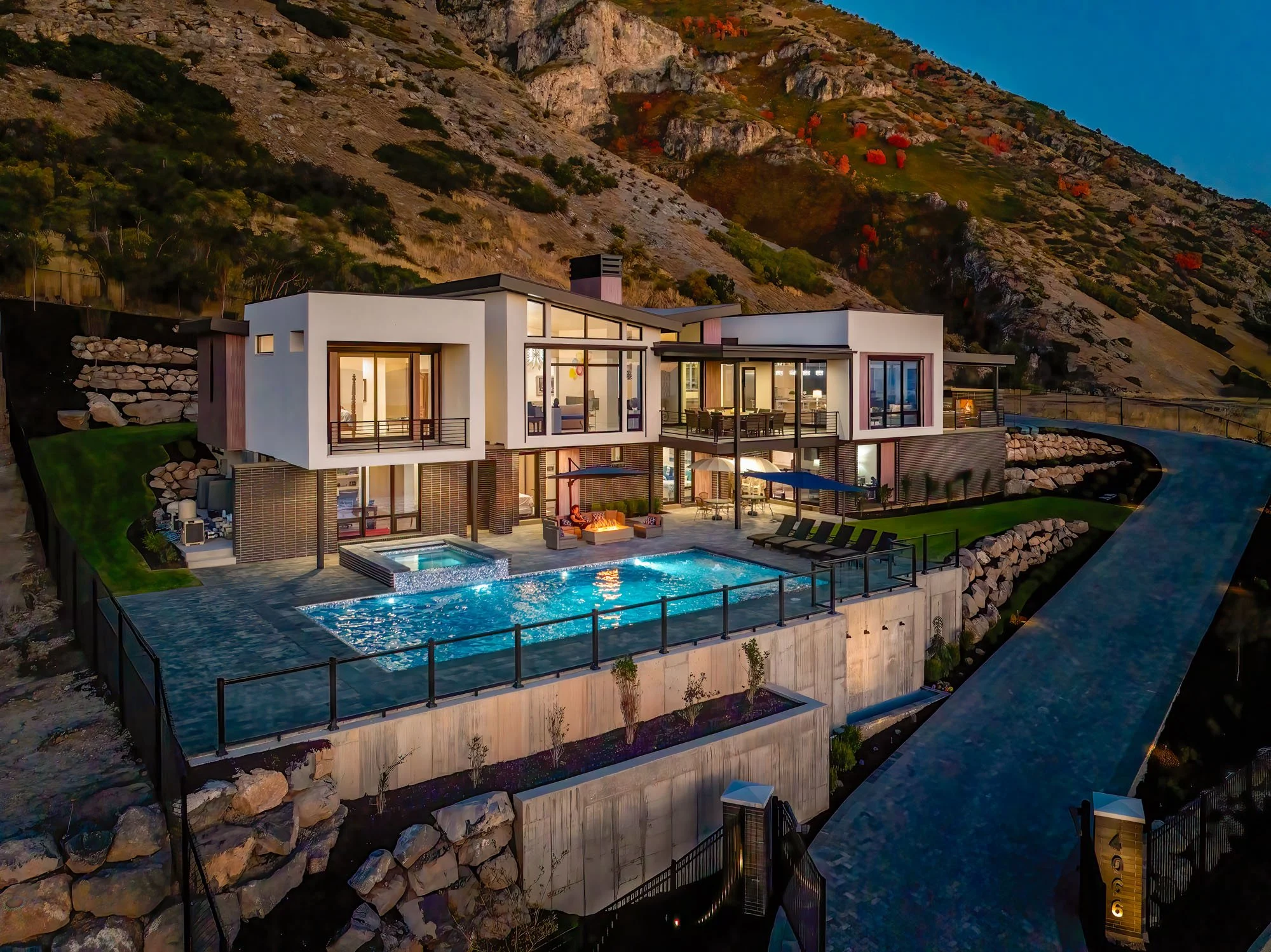 Luxury modern house situated on a hillside at dusk, featuring large glass windows and multiple balconies. The house includes a swimming pool and spa area with outdoor seating, surrounded by rocks and landscaping, with a paved driveway leading up to the home.