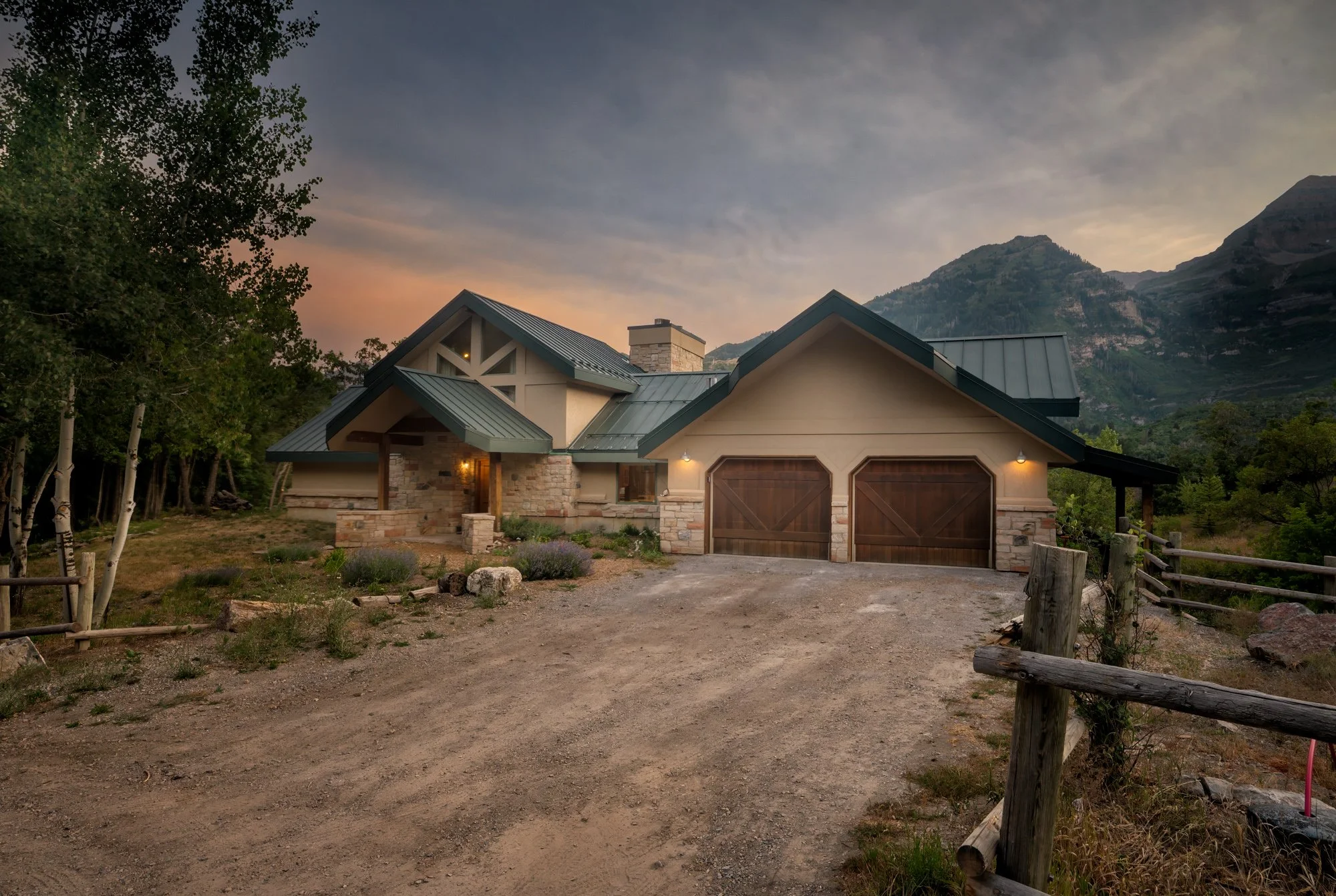 A house with a natural stone exterior, dark brown garage doors, and a metal roof, set against a mountainous backdrop at sunset.