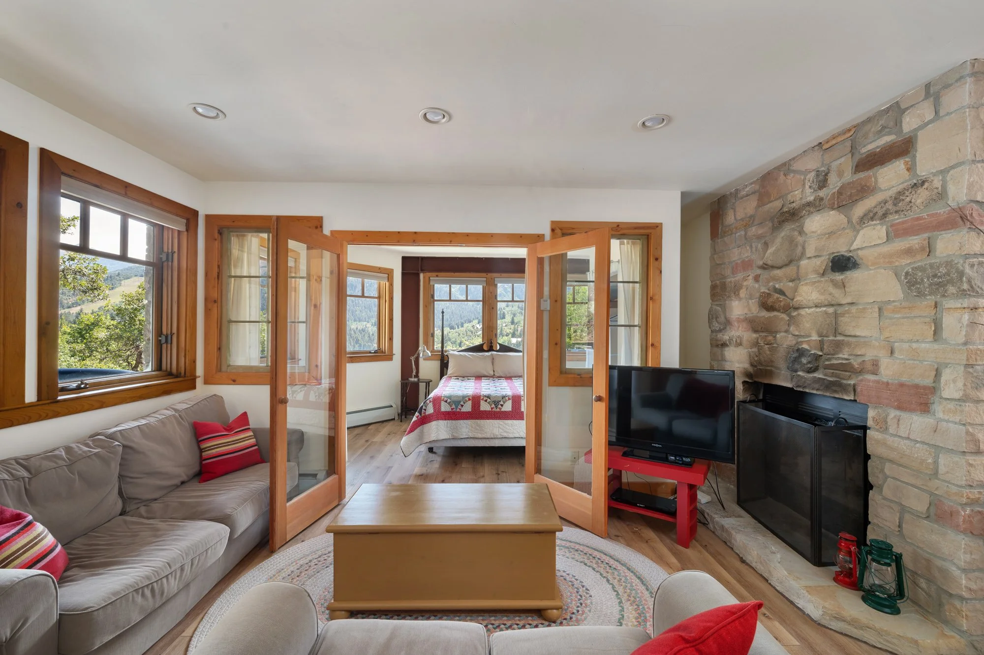 Living room with beige sofa, red and beige striped pillows, wooden coffee table, TV on a red stand, brick fireplace, and open bedroom with bed and windows showing scenic landscape outside.