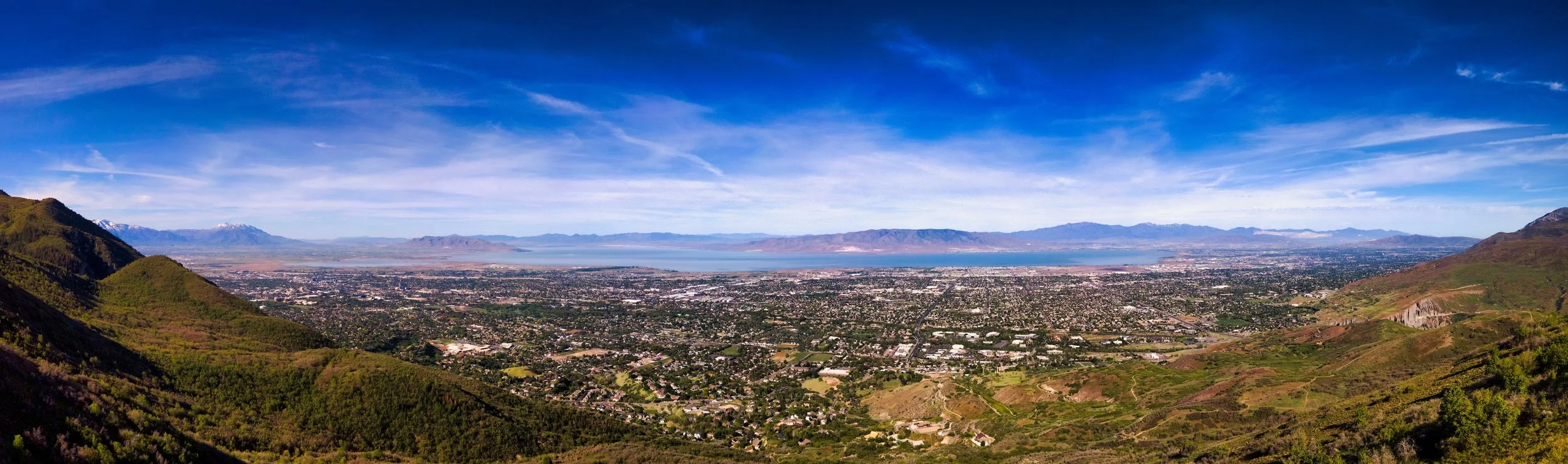A panoramic view of a city from a hillside with mountains and a lake in the distance under a blue sky with wispy clouds.