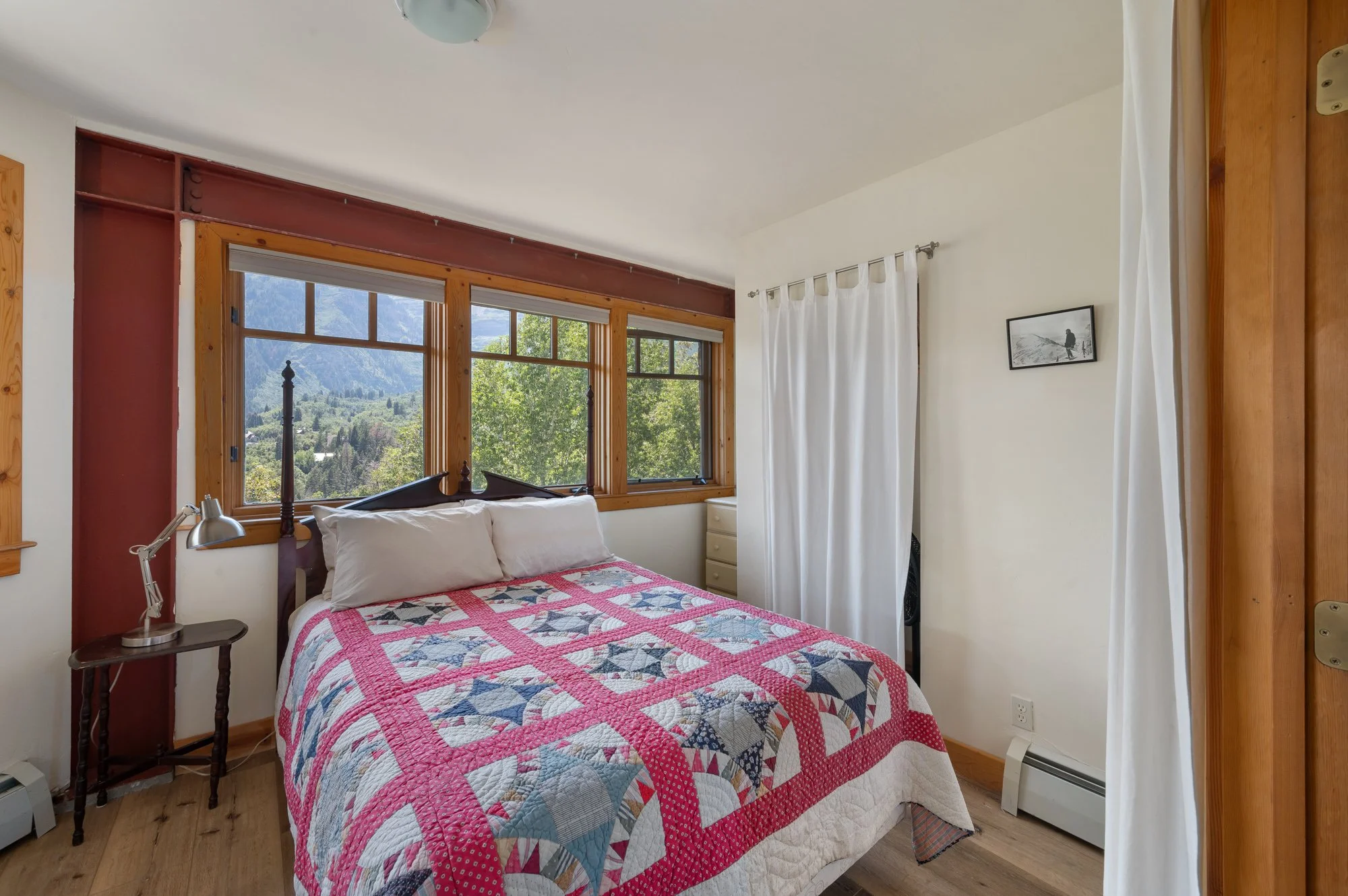 Cozy bedroom with large windows revealing mountain view, bed with quilt, white pillows, and wooden furniture.