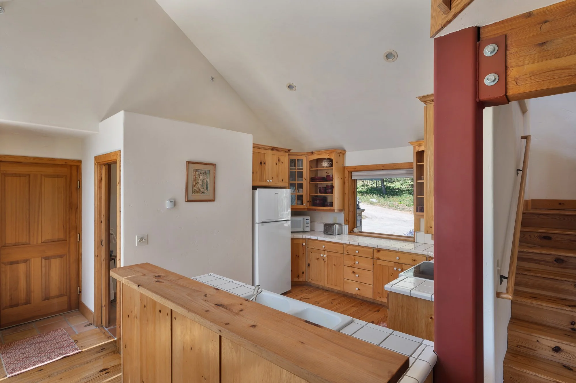 Kitchen with wooden cabinets, white refrigerator, microwave, toaster, window showing outdoor greenery, staircase, and white tiled counter.