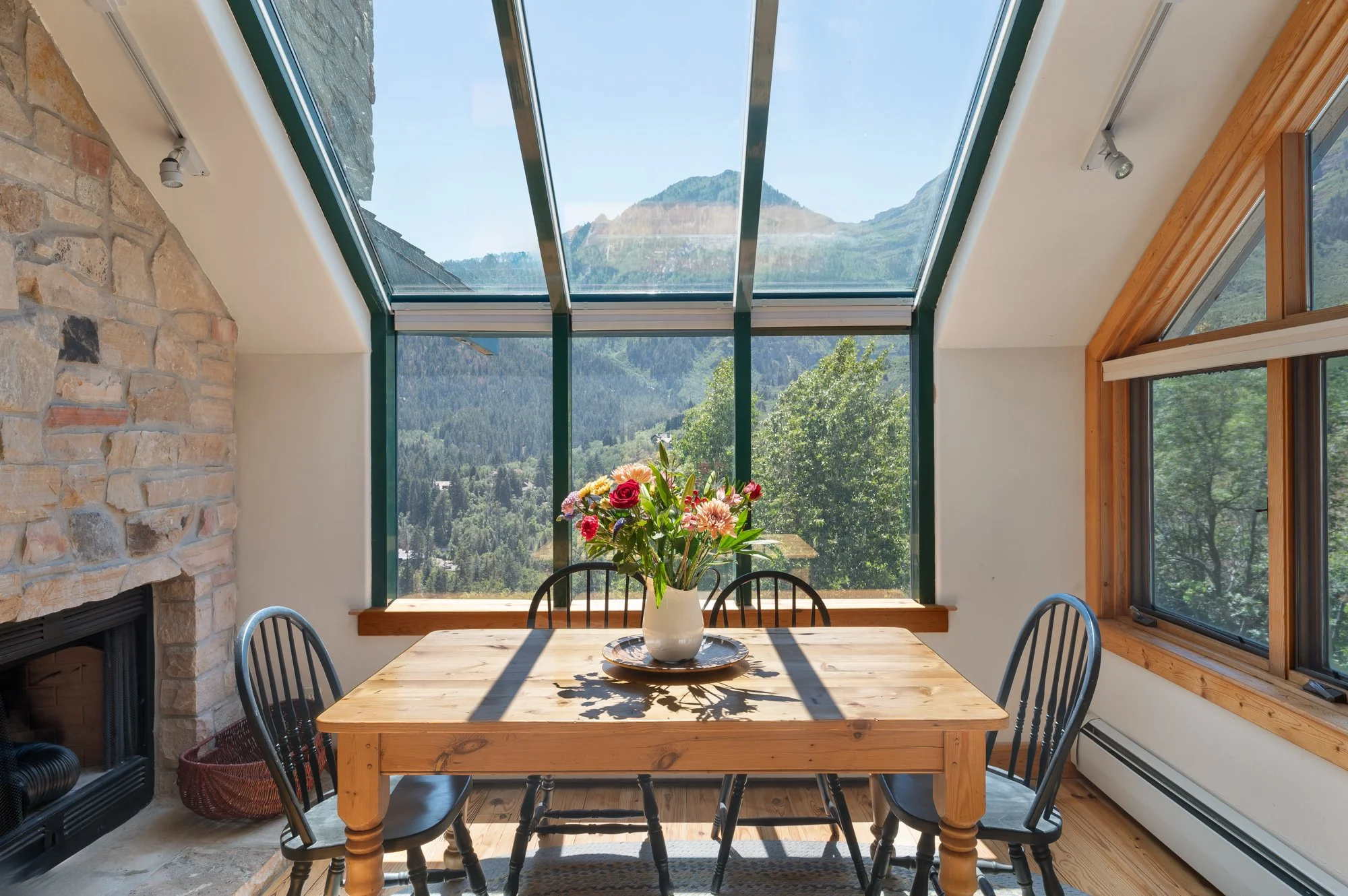 Dining area with a wooden table and black chairs, a vase of colorful flowers, large windows showing a mountain view, stone fireplace on the left, and wooden trim around the windows.