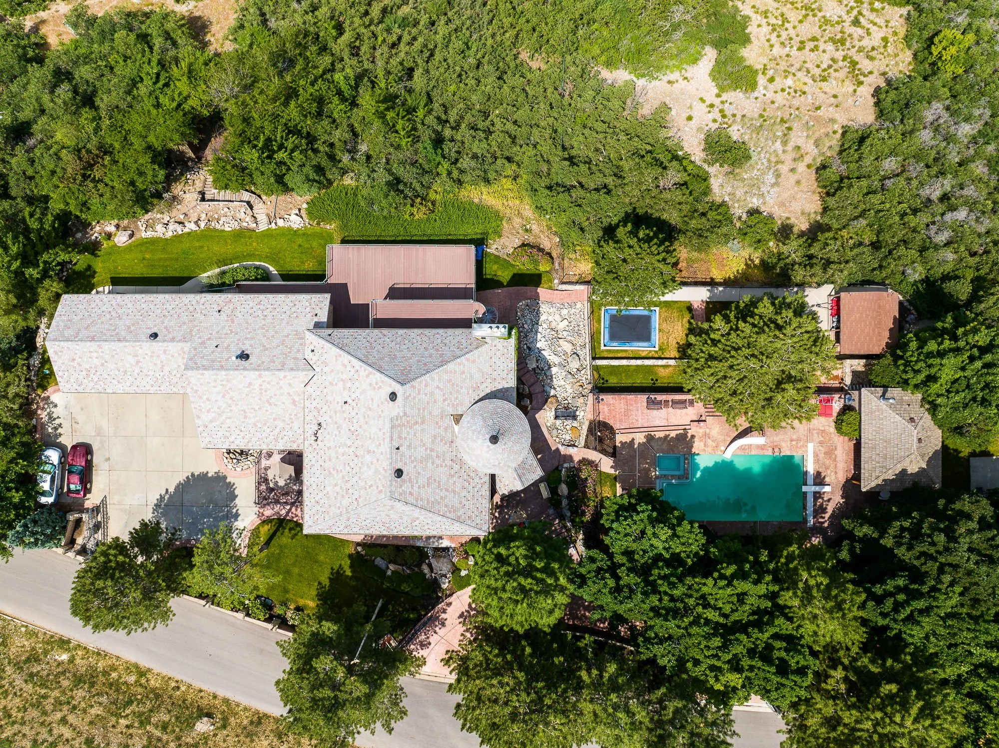 An aerial view of a house with a large roof, surrounded by trees, a driveway with cars, a swimming pool, and a fenced yard with lush green grass.