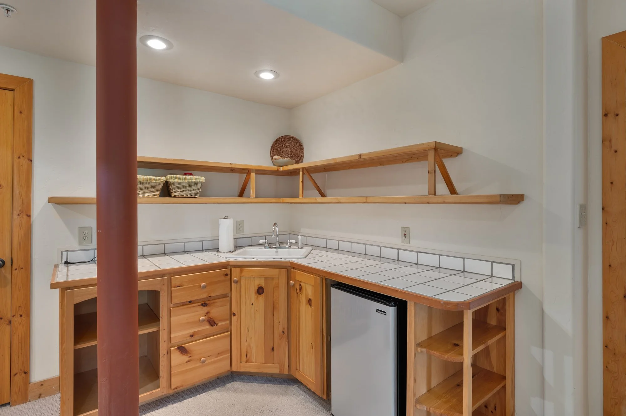 Small kitchen area with wooden cabinets, white tiled countertop, small refrigerator, and open shelving. The area has a white wall and ceiling with built-in overhead lights.
