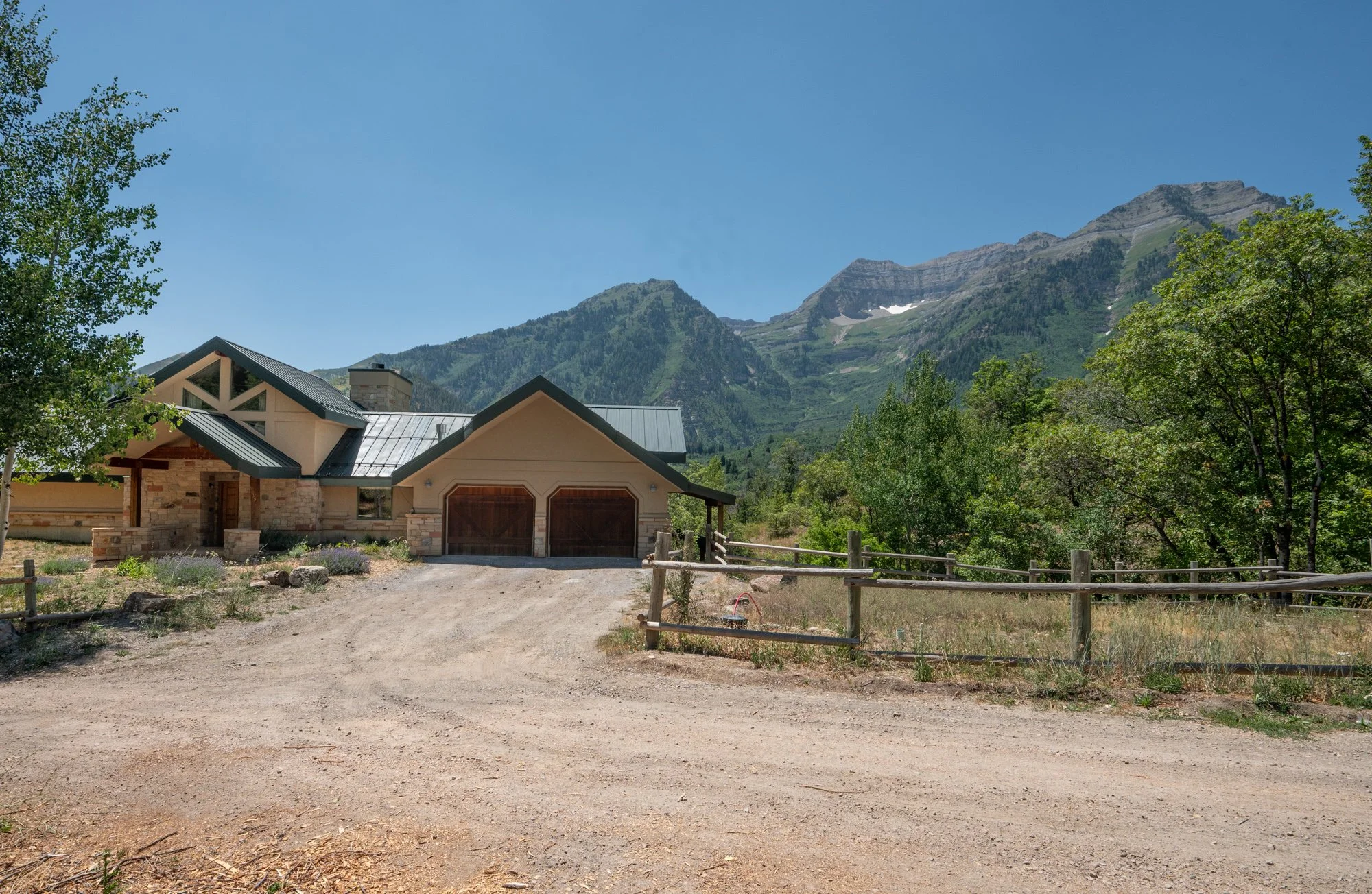 A house with stone and beige siding, garage doors, and a metal roof, set against green trees and mountains under a clear blue sky.