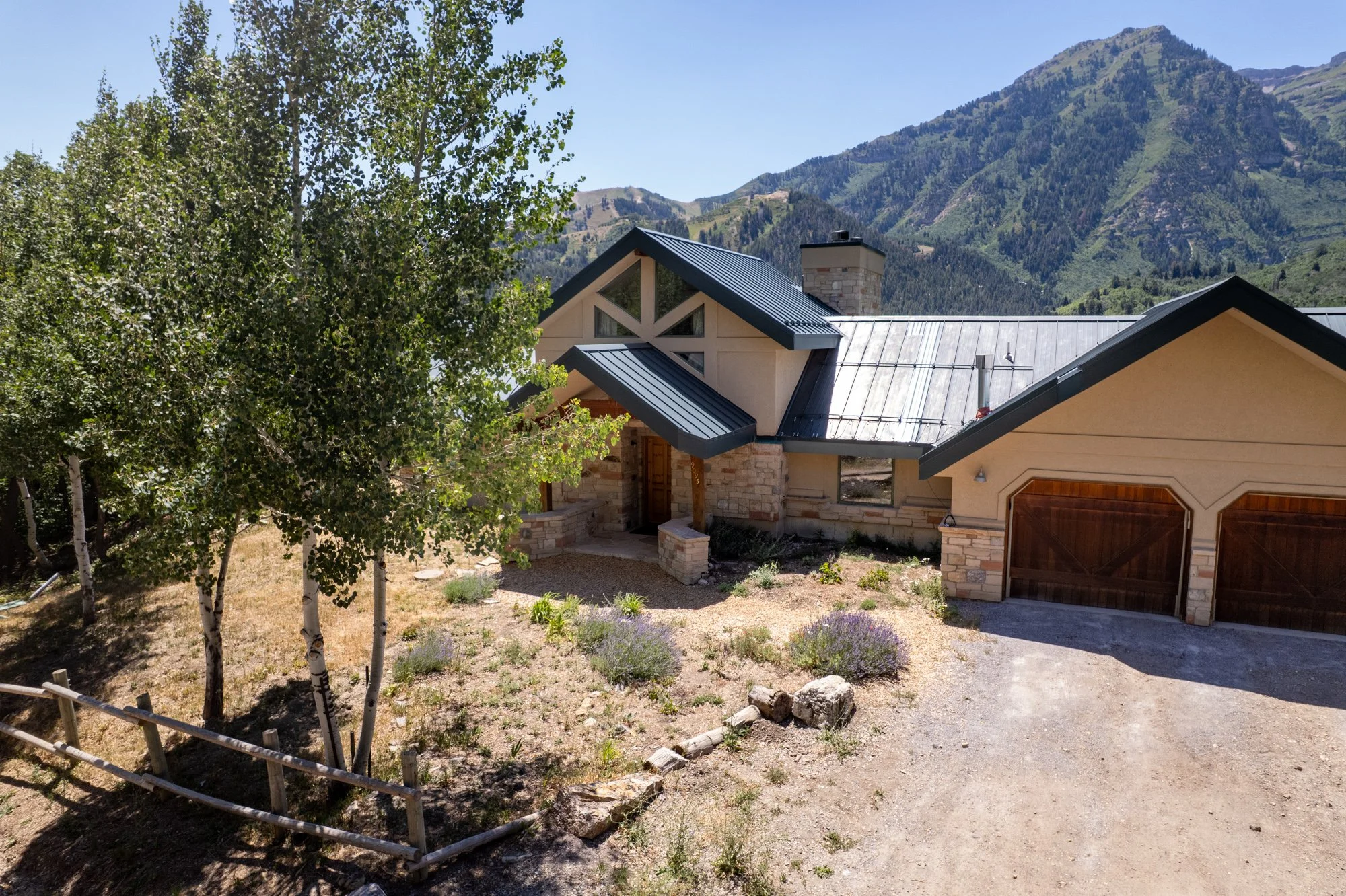 A house with a metal roof in a mountainous landscape, surrounded by trees and sparse landscaping.