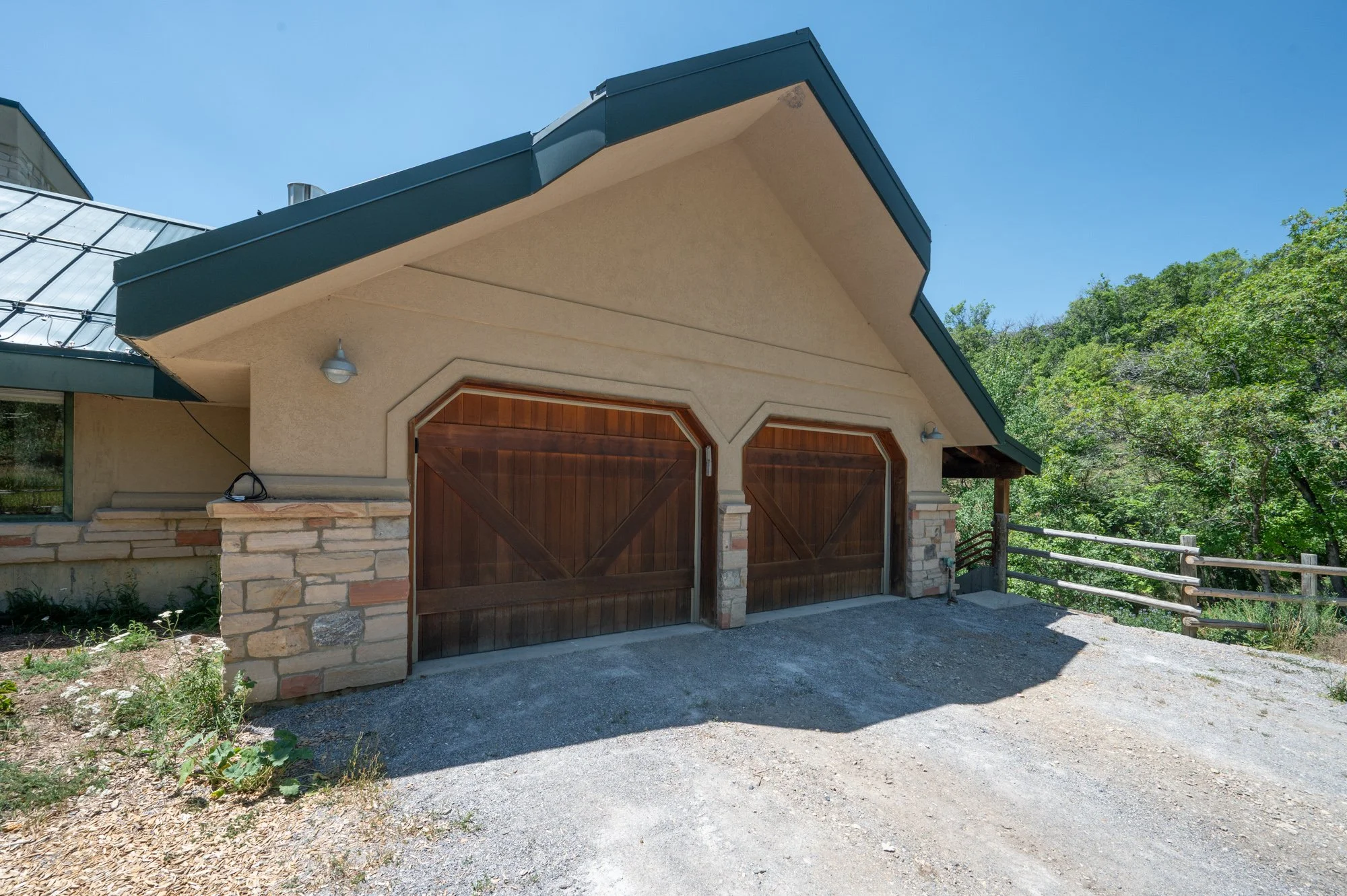 A house's garage with two wooden garage doors, stone pillars, and a green roof, located in a rural area with trees and a gravel driveway.