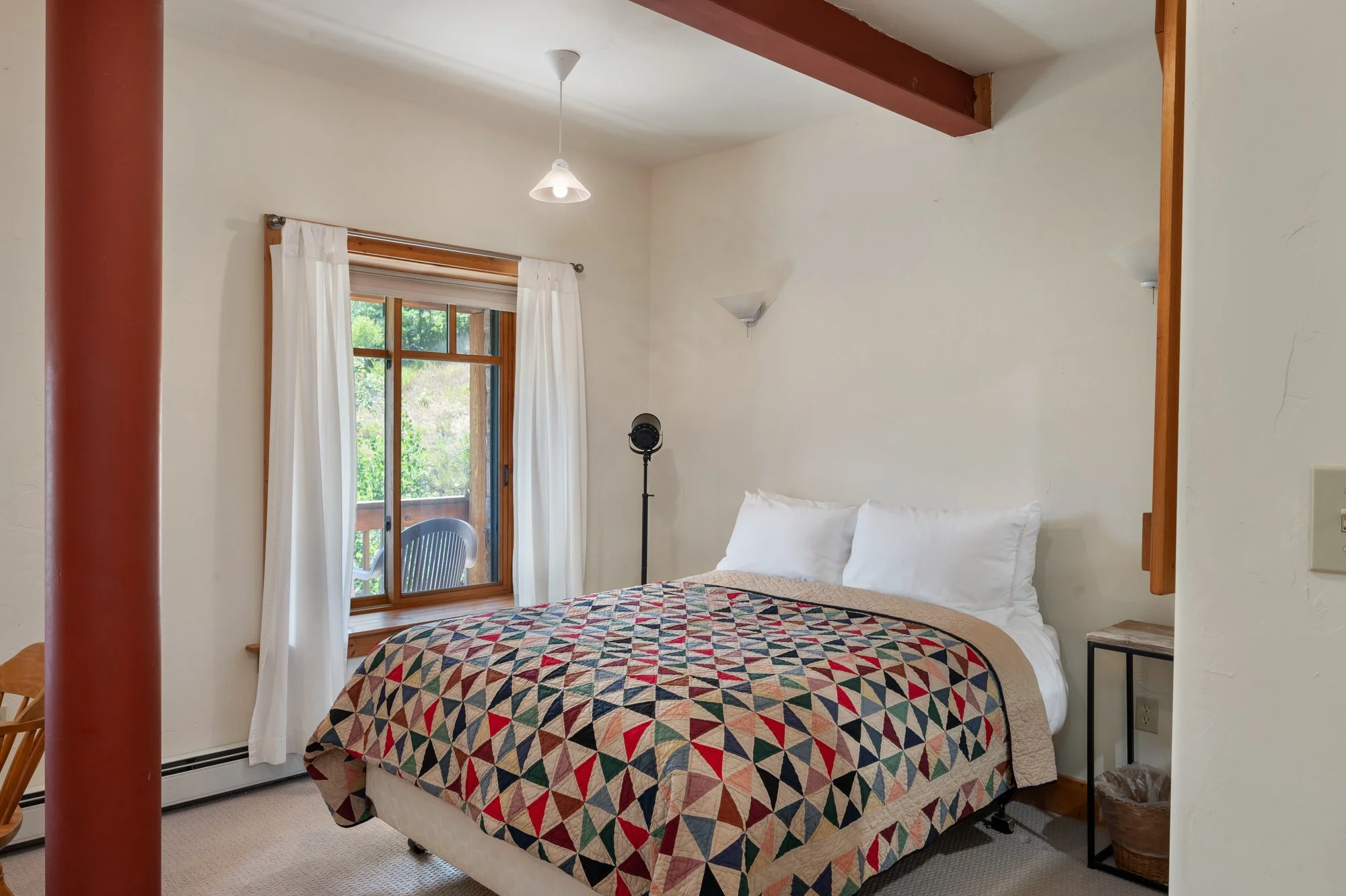 A cozy bedroom with a bed covered in a colorful geometric quilt, two white pillows, a side table, and a window with white curtains showing outdoor greenery.