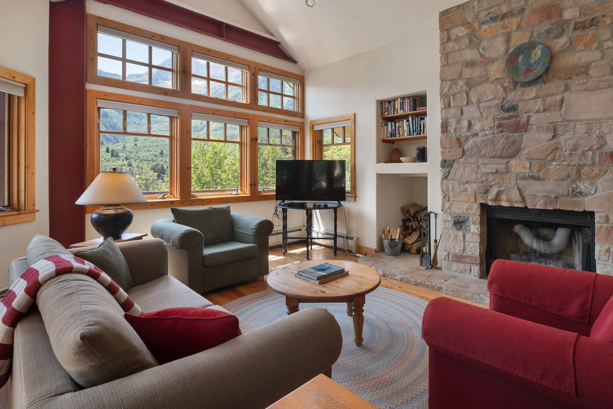 Living room with large windows showing mountain view, stone fireplace, sofa, armchair, coffee table, bookshelf, and flat-screen TV.