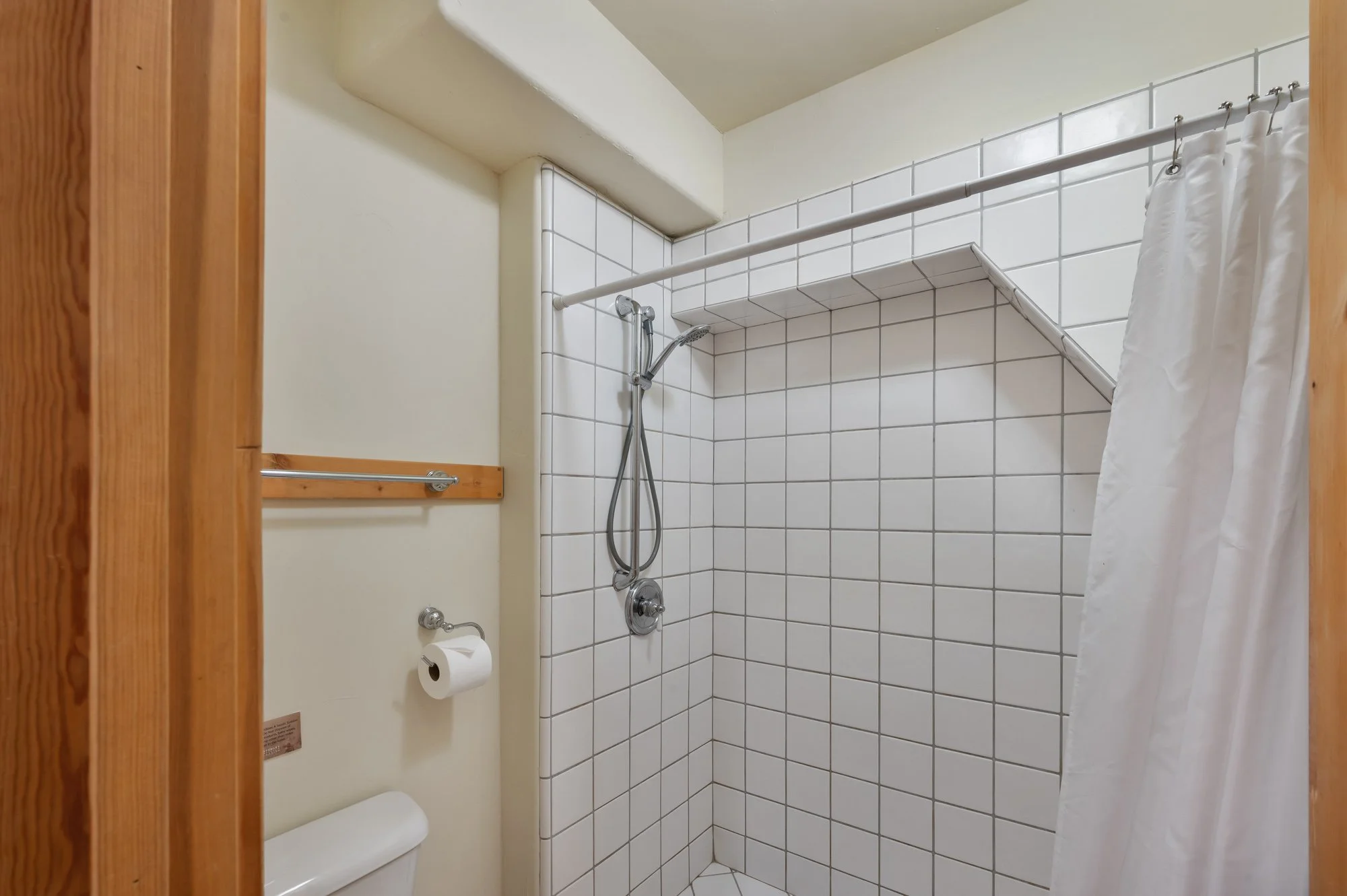 Small bathroom with a tiled shower, white curtain, toilet, toilet paper, and wooden trim.