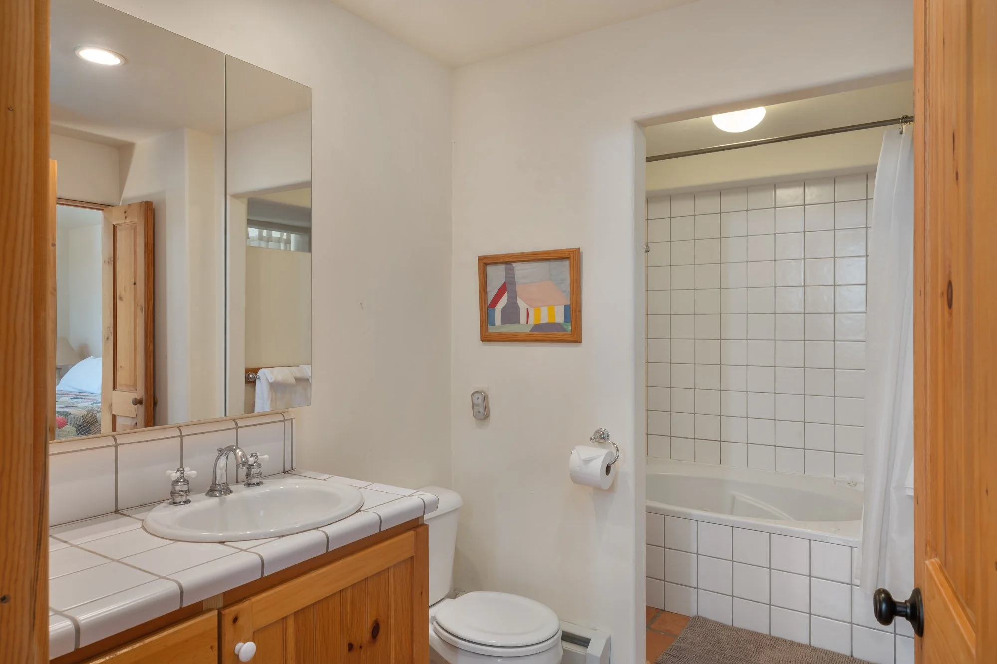 Bathroom with white walls and tile features, including a sink with a mirror, toilet, and a bathtub with a shower curtain, wood accents, and a small wall art.