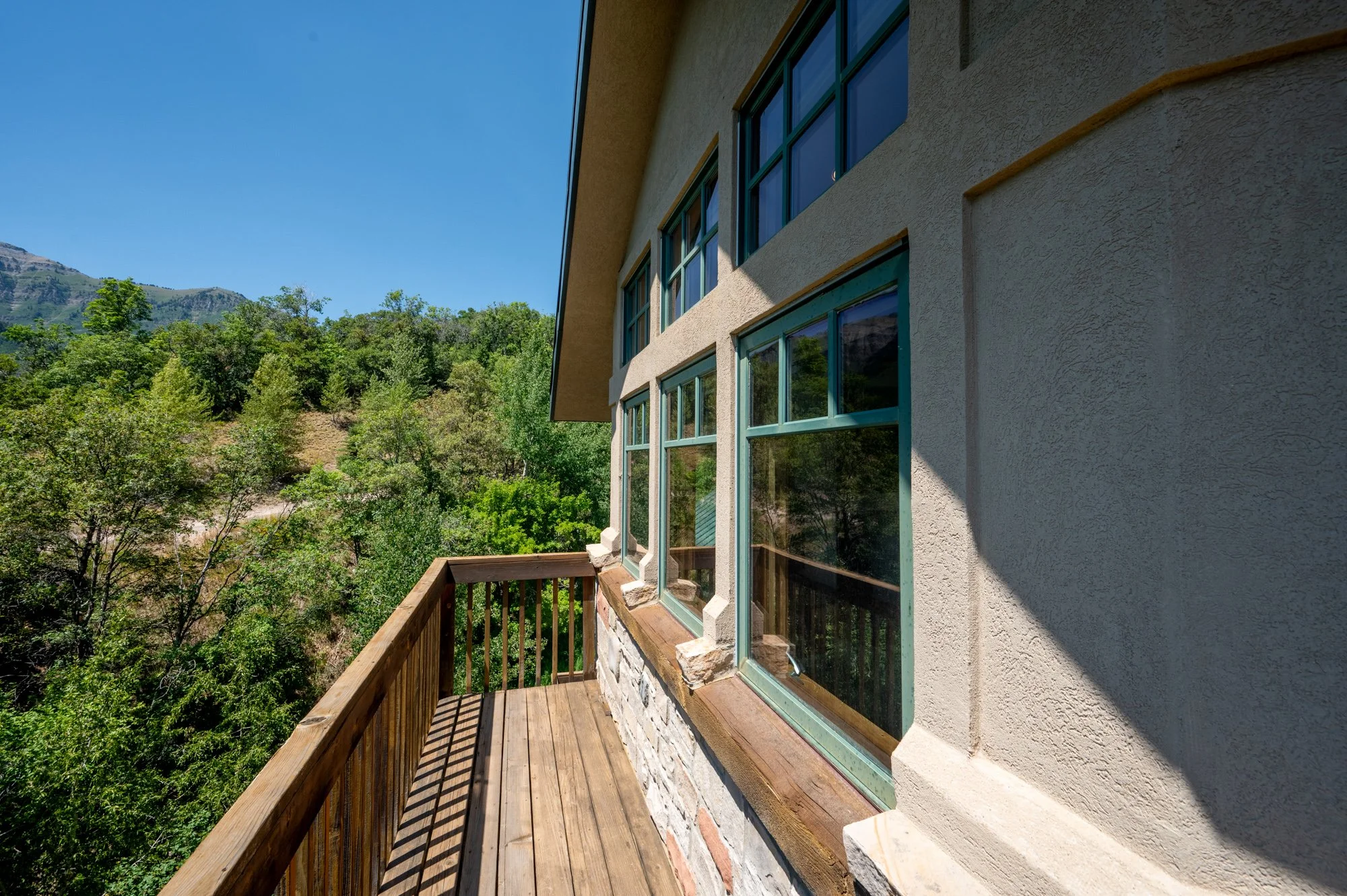 Wooden balcony with railing attached to a beige stucco house, overlooking a landscape with green trees and mountains under a clear blue sky.