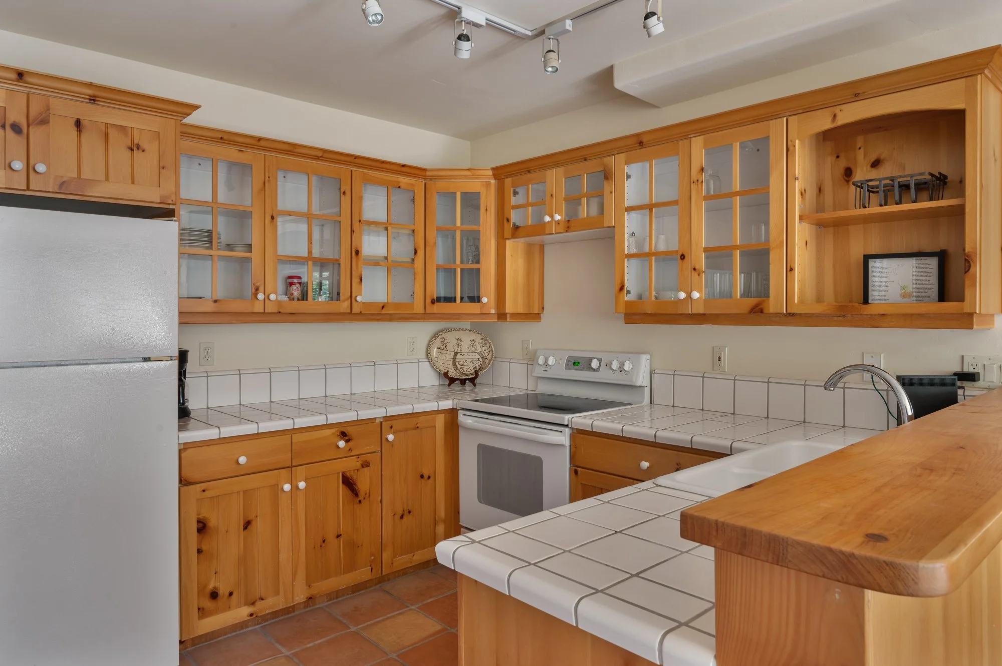 Kitchen with wooden cabinets, white tiled countertops, white stove, refrigerator, and a double sink, with terracotta tile flooring.