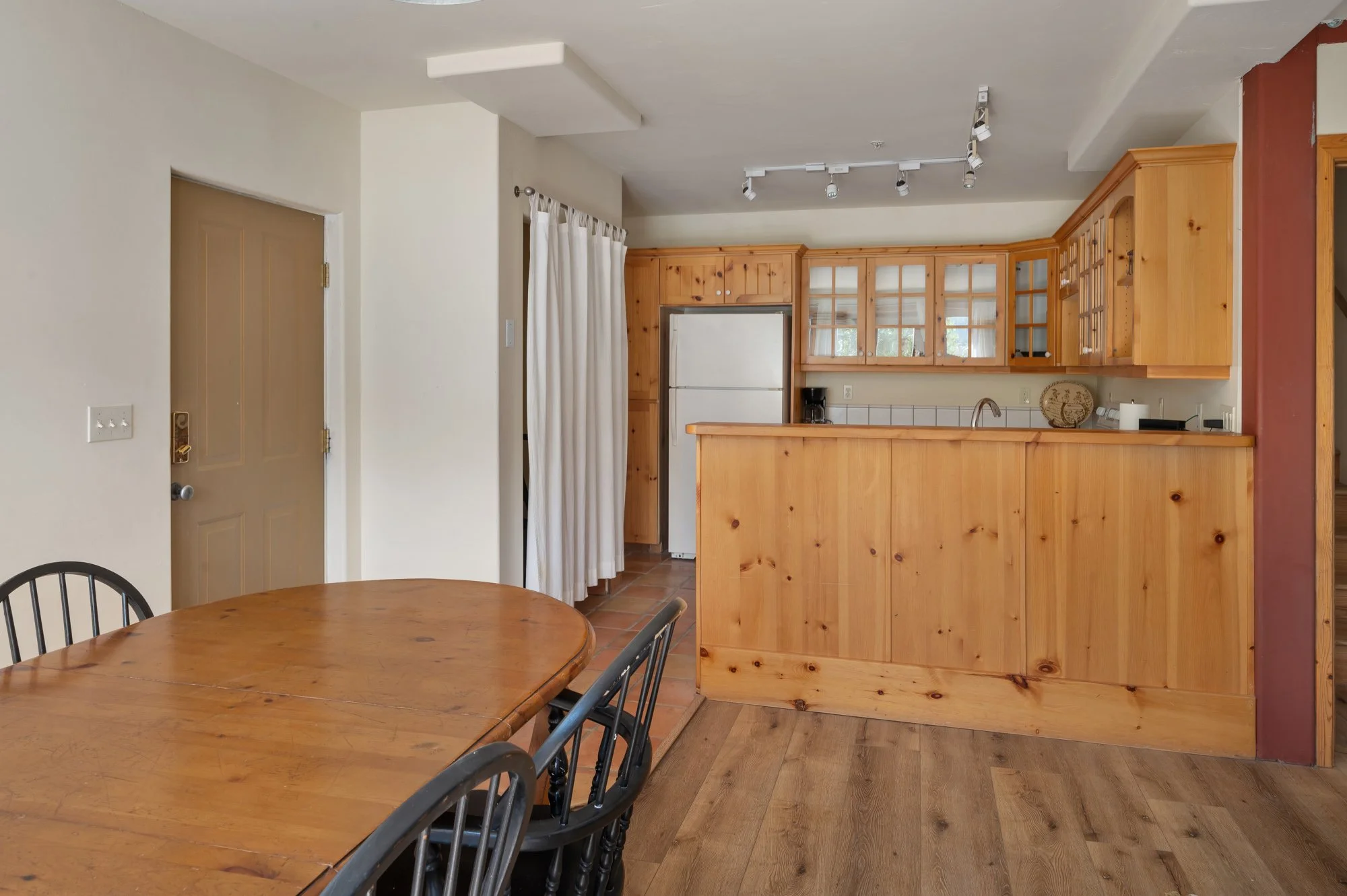 Interior view of a kitchen with wooden cabinets, a white refrigerator, and a breakfast bar. There is a dining table with black chairs in the foreground. White curtains cover a doorway, and the floor is wood with a reddish accent wall.