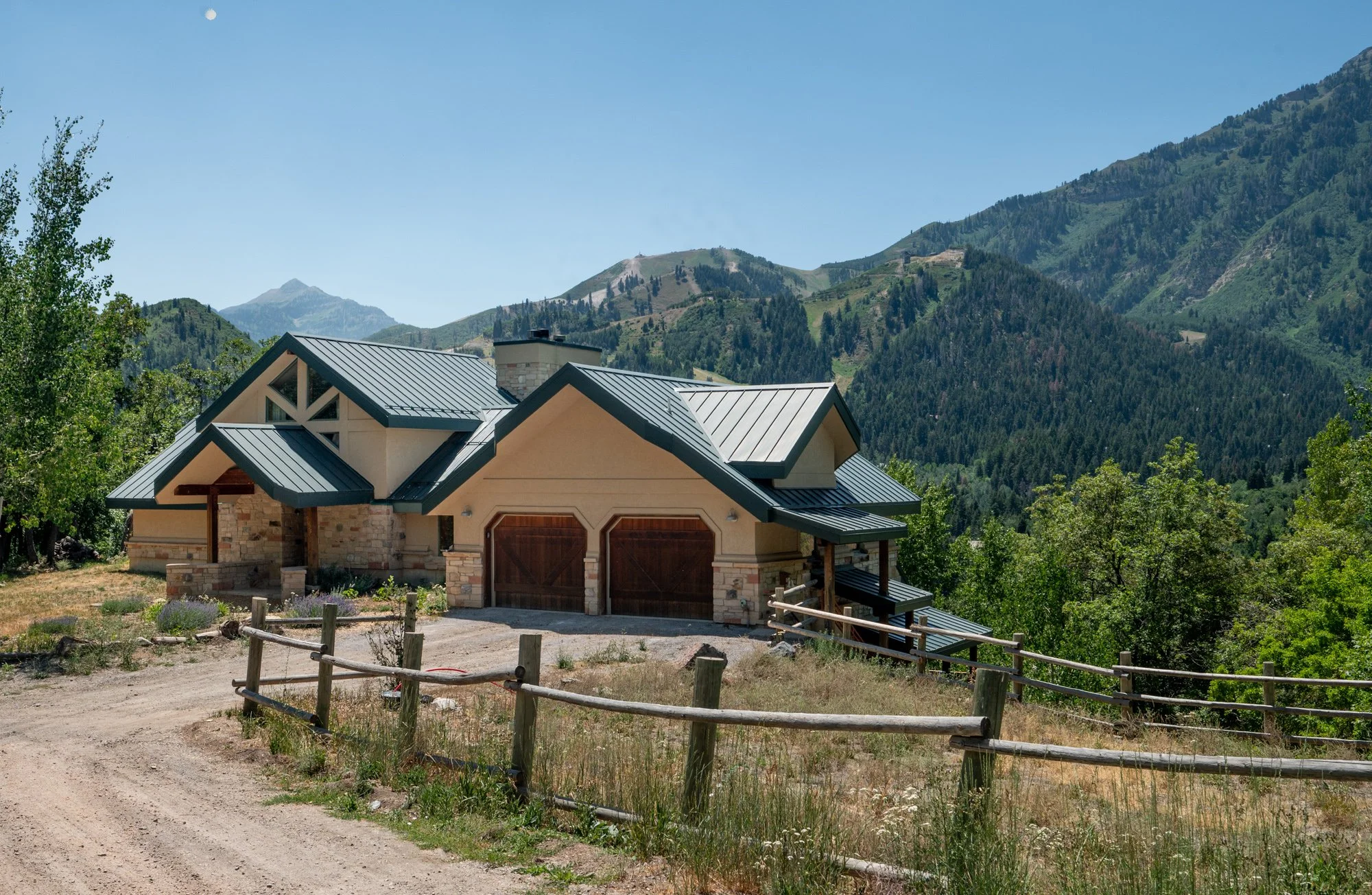 A modern house with a metal roof, stone accents, surrounded by green trees and mountains in the background, on a dirt road.