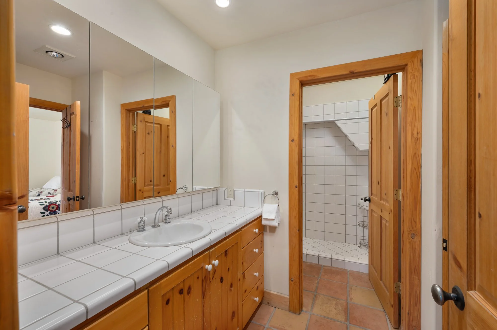 Bathroom with white tiled countertop and wooden cabinetry, mirror, and a doorway leading to a tiled shower with a built-in shelf.
