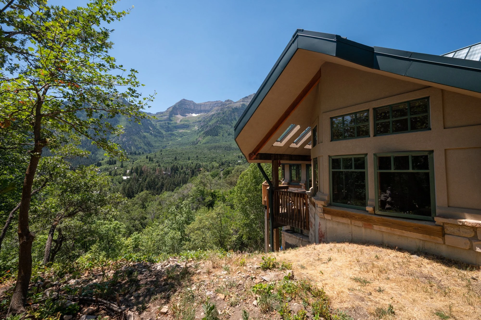 A house on a hillside with large windows and a balcony, surrounded by trees, with a forested mountain in the background under a clear blue sky.