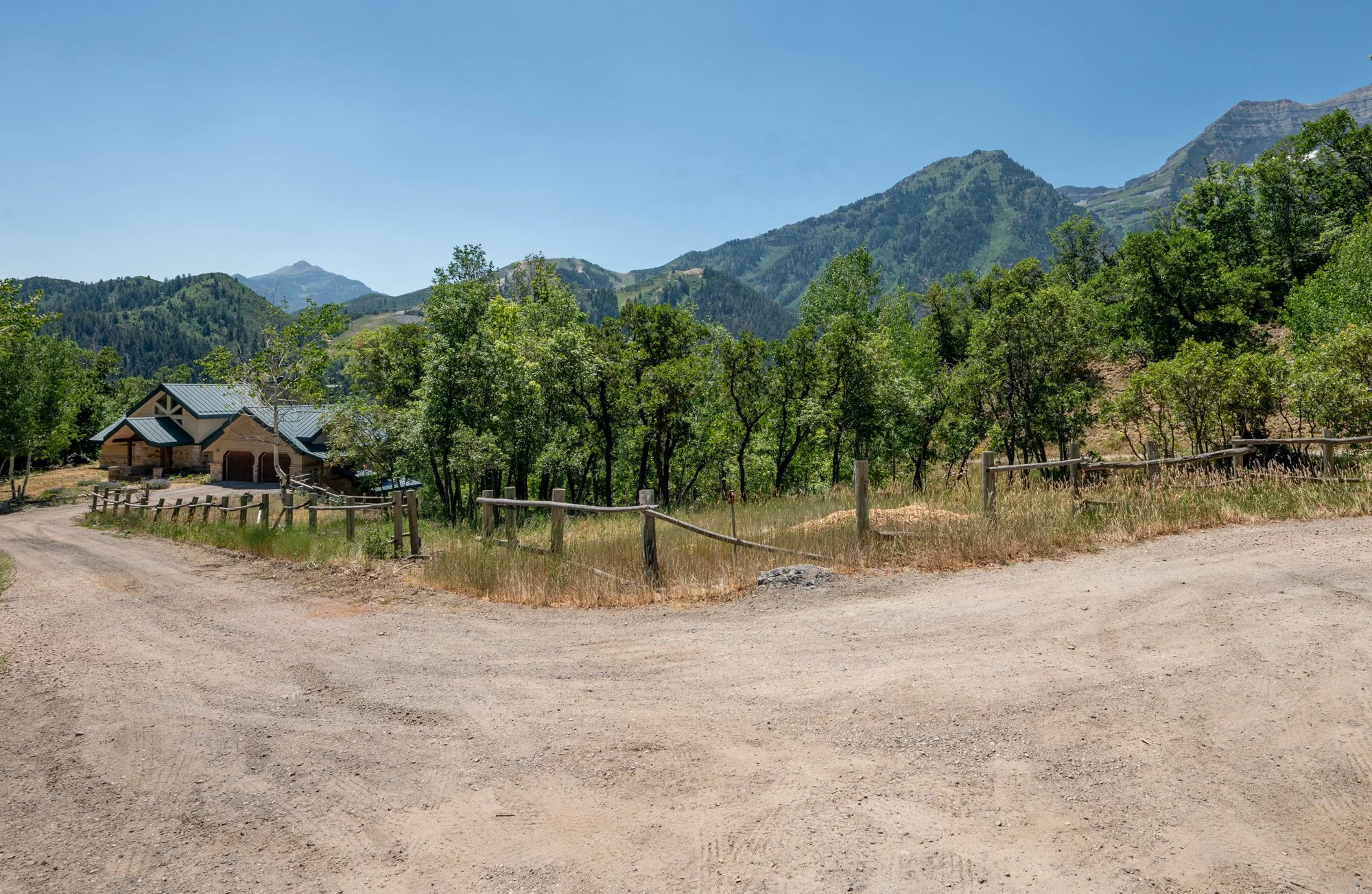 A house with a gray roof is surrounded by green trees, set against a mountainous landscape under a clear blue sky, with a dirt road leading to it.