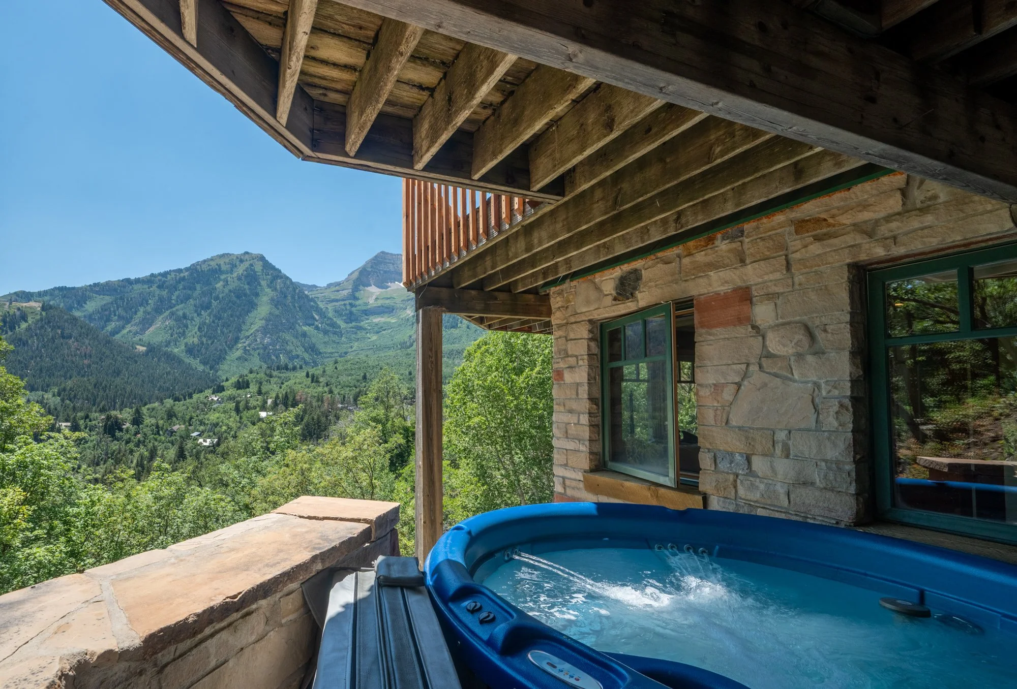 A steaming hot tub on a rustic balcony with stone walls and wooden beams, overlooking green mountains and a forested landscape.