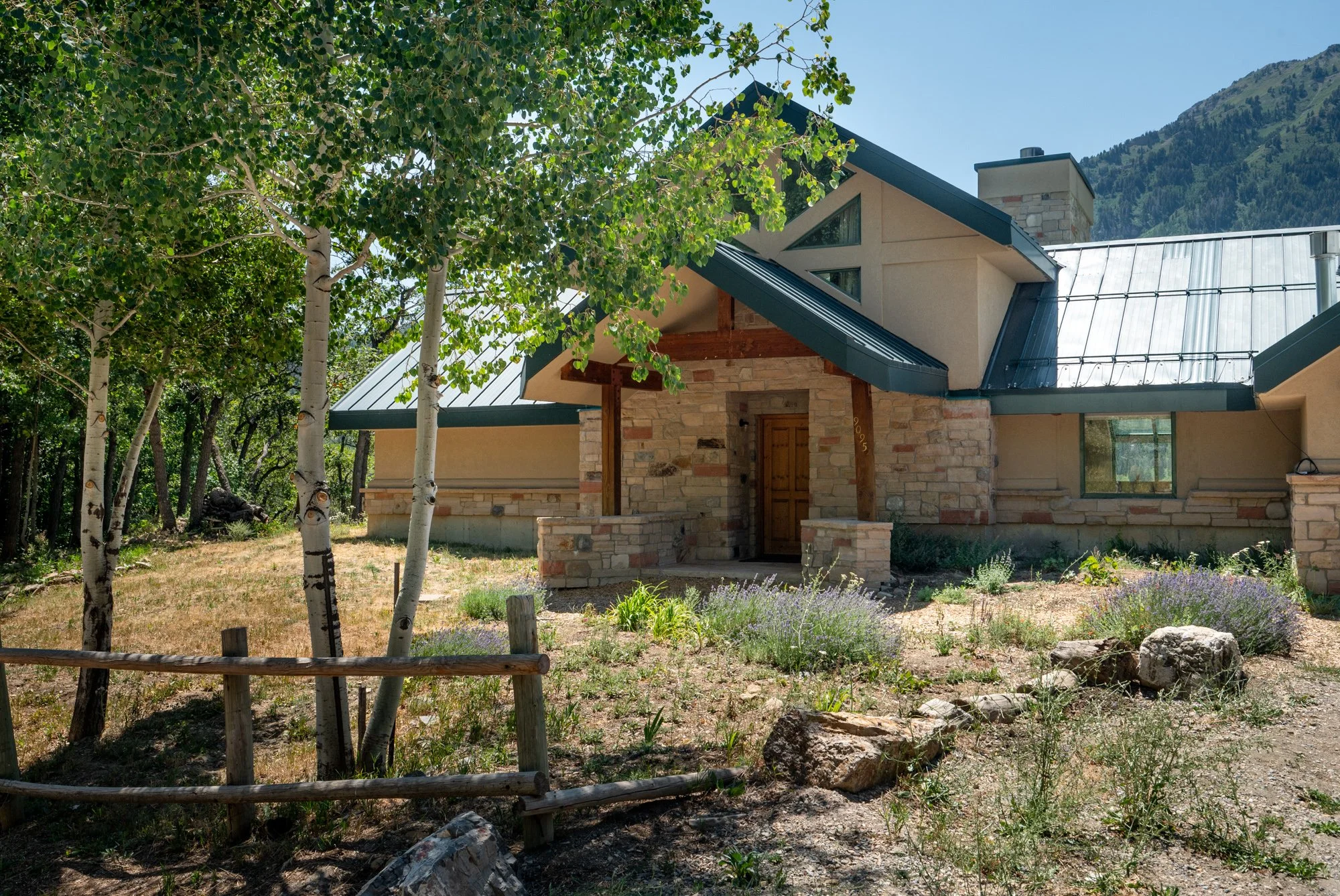 A house with a stone and wood exterior, metal roof, and a small porch entrance, surrounded by trees and rocks, with a mountain in the background.