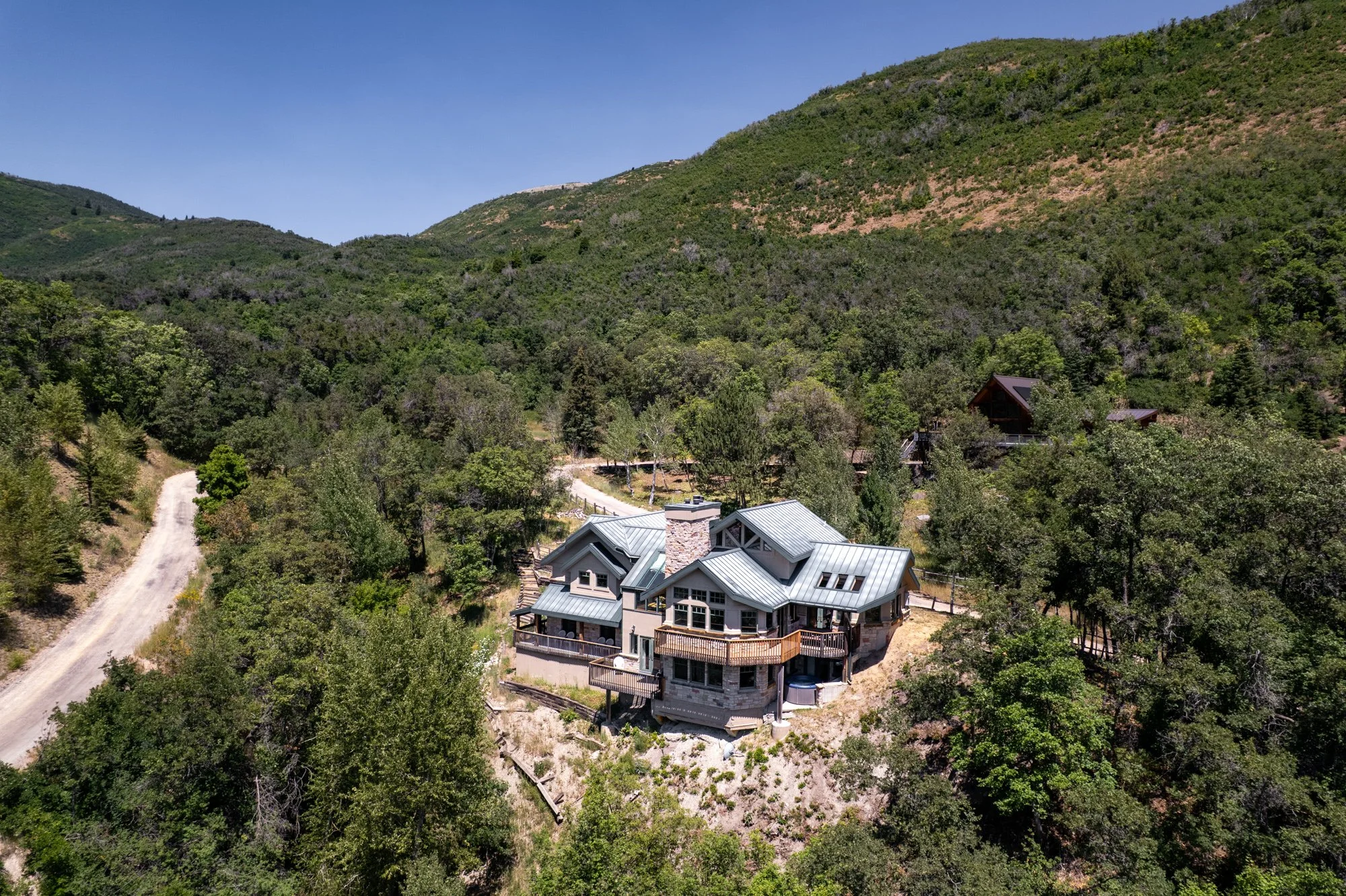 A large house with metal roofing nestled among trees on a hillside with mountains in the background.