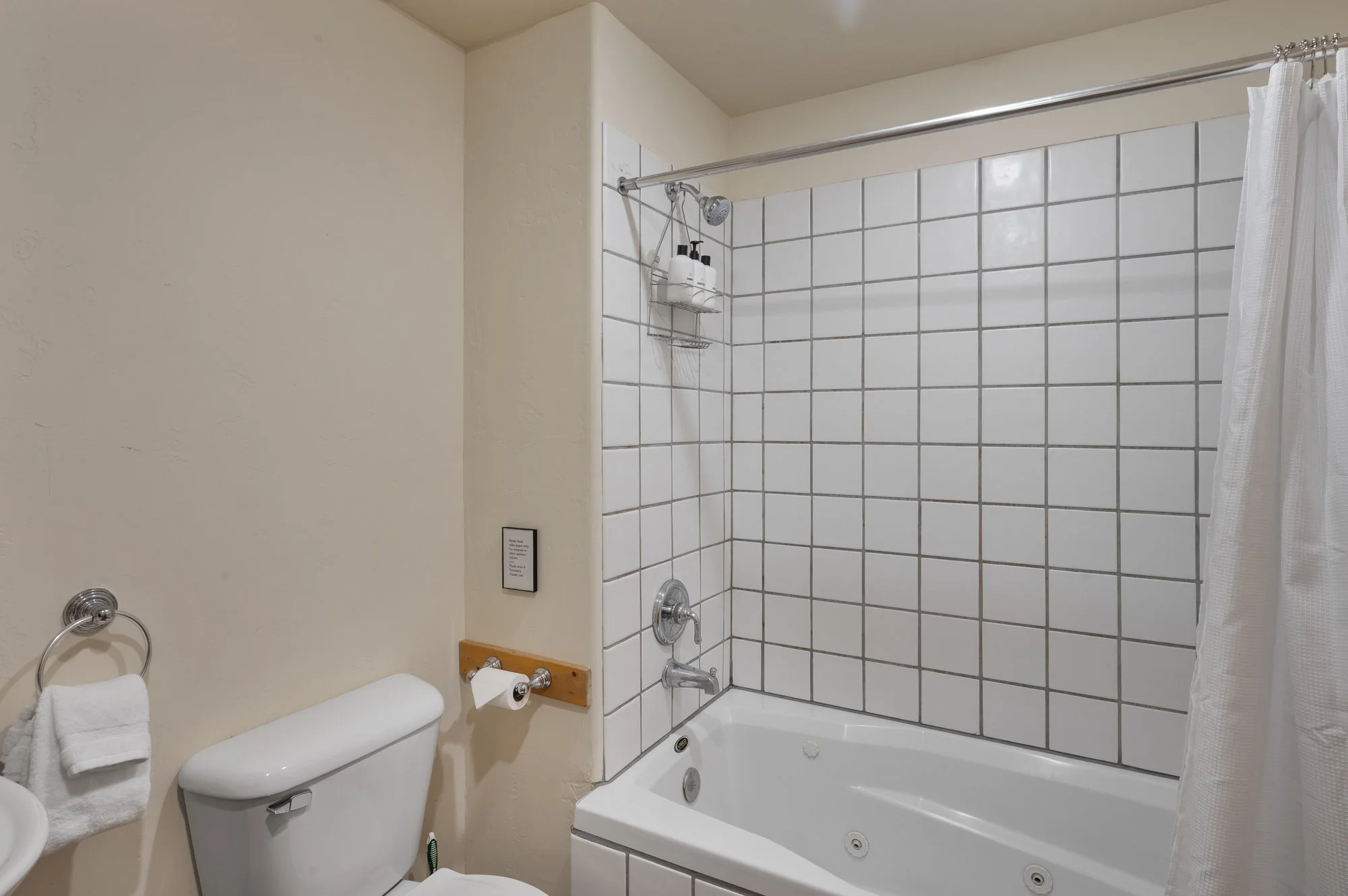 Bathroom with beige walls, a toilet with a towel hanging on a silver ring, and a bathtub with white square tiles on the walls surrounding it. The bathtub has a showerhead, faucet, and a shower caddy holding soap and shampoo. A shower curtain partiall