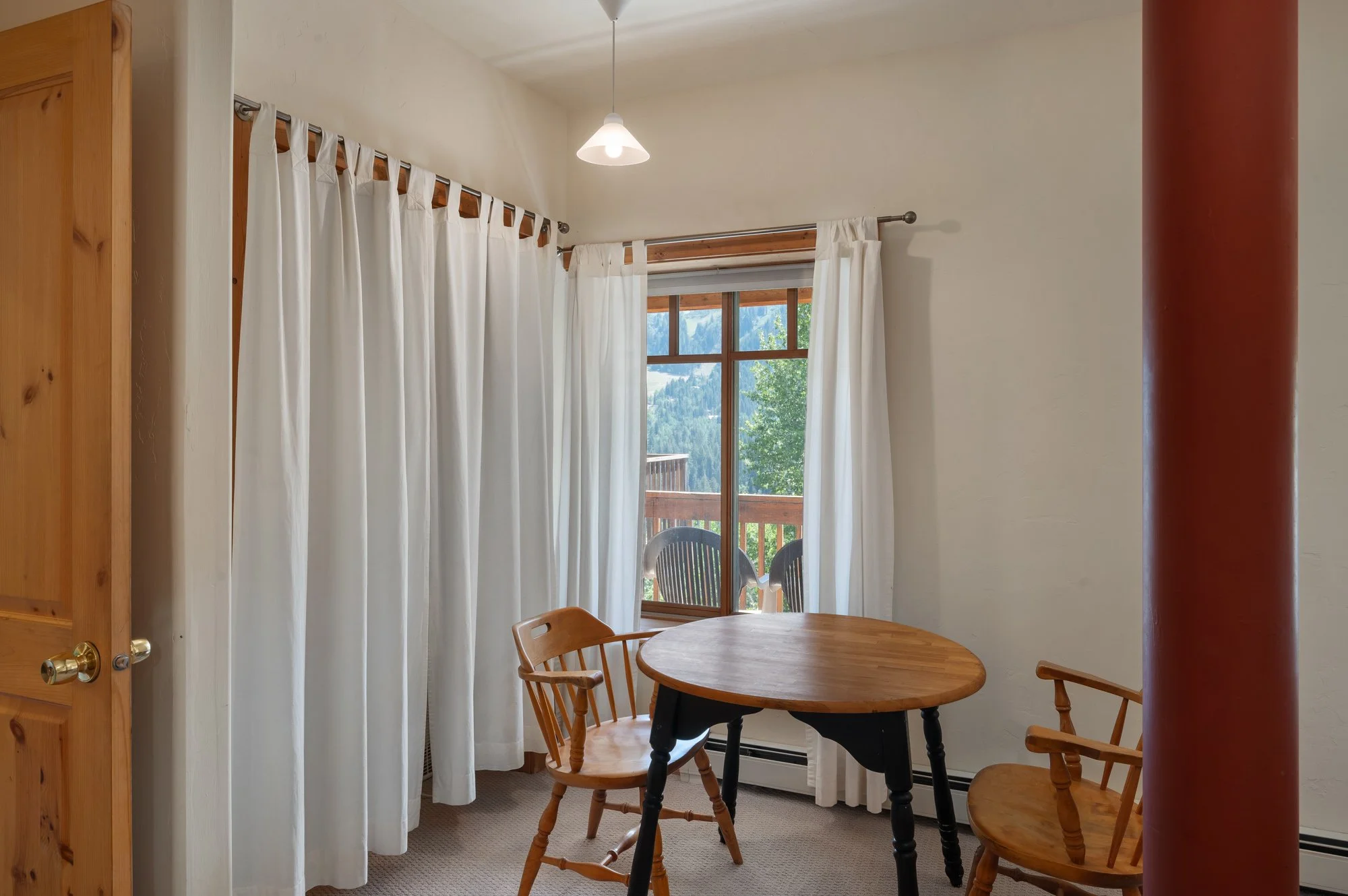 Dining area with a wooden table and chairs, large window with white curtains, view of an outdoor balcony with chairs and trees, neutral-colored walls, and ceiling light fixture.