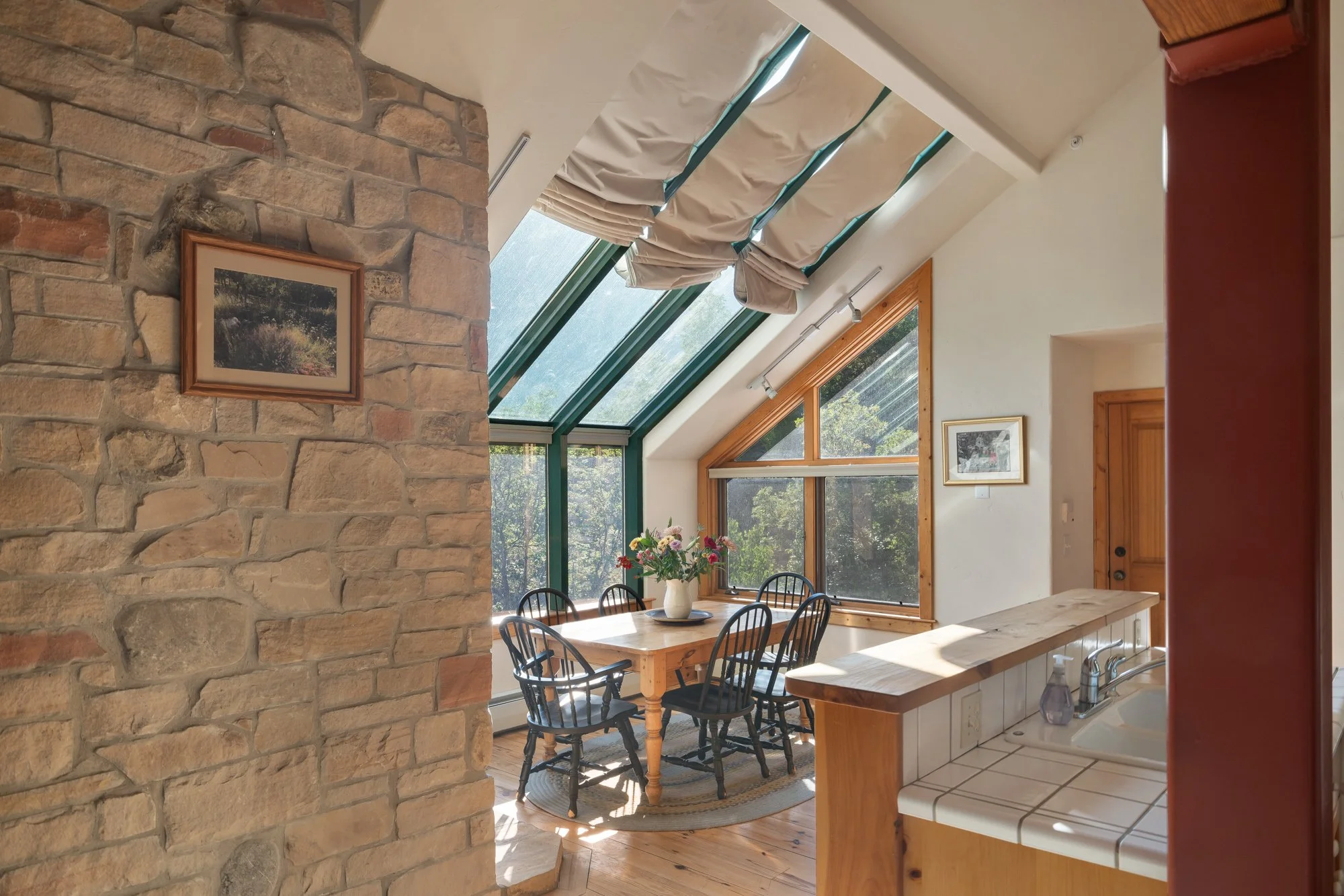 Dining area with a wooden table, six black chairs, a vase with flowers, large windows, and a sloped glass ceiling, adjacent to a kitchen with tiled countertop and a sink. A framed picture hangs on the stone wall.