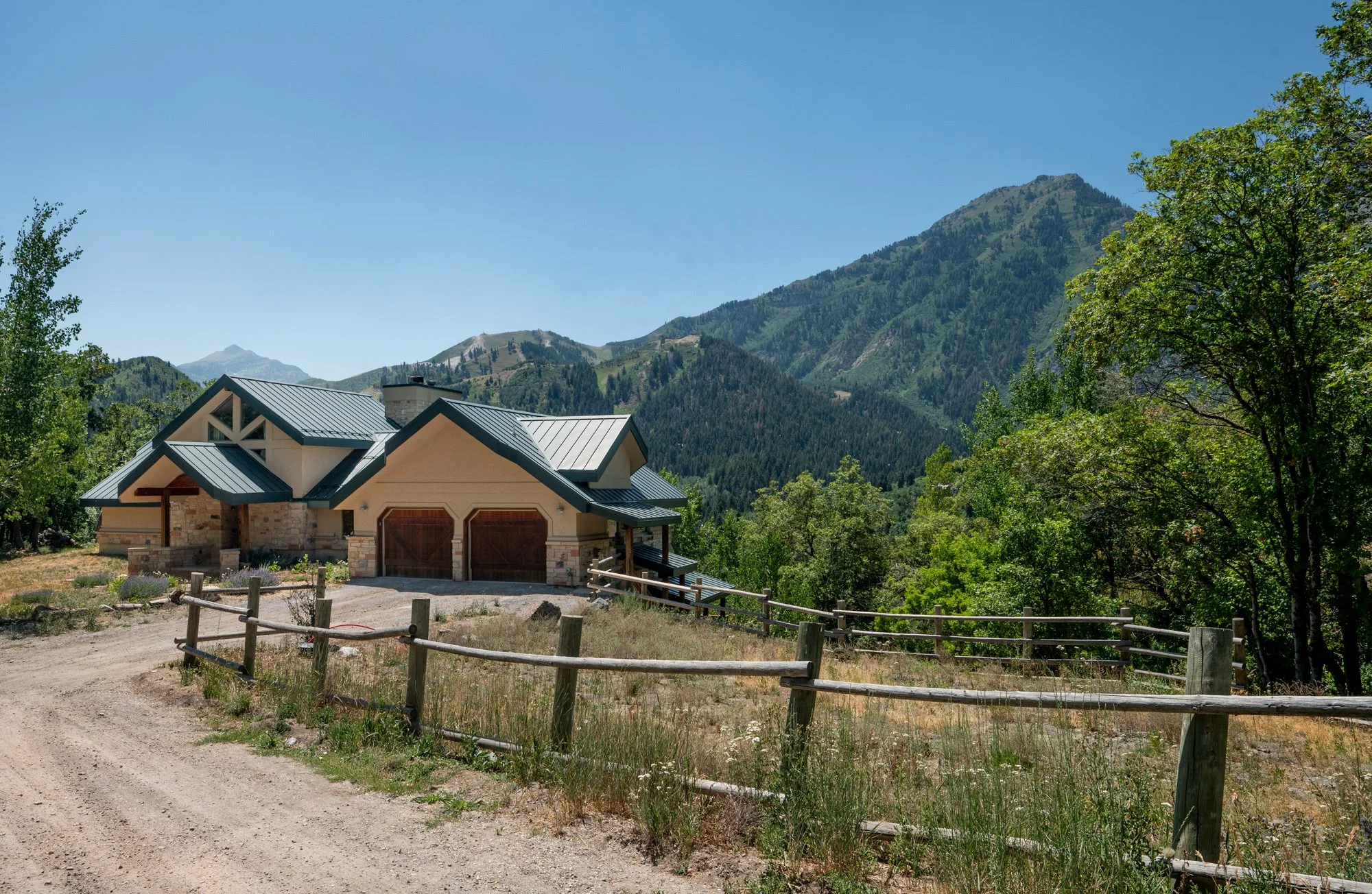 A house with a metal roof and wood garage doors is situated on a dirt road surrounded by green trees and mountains in the background.