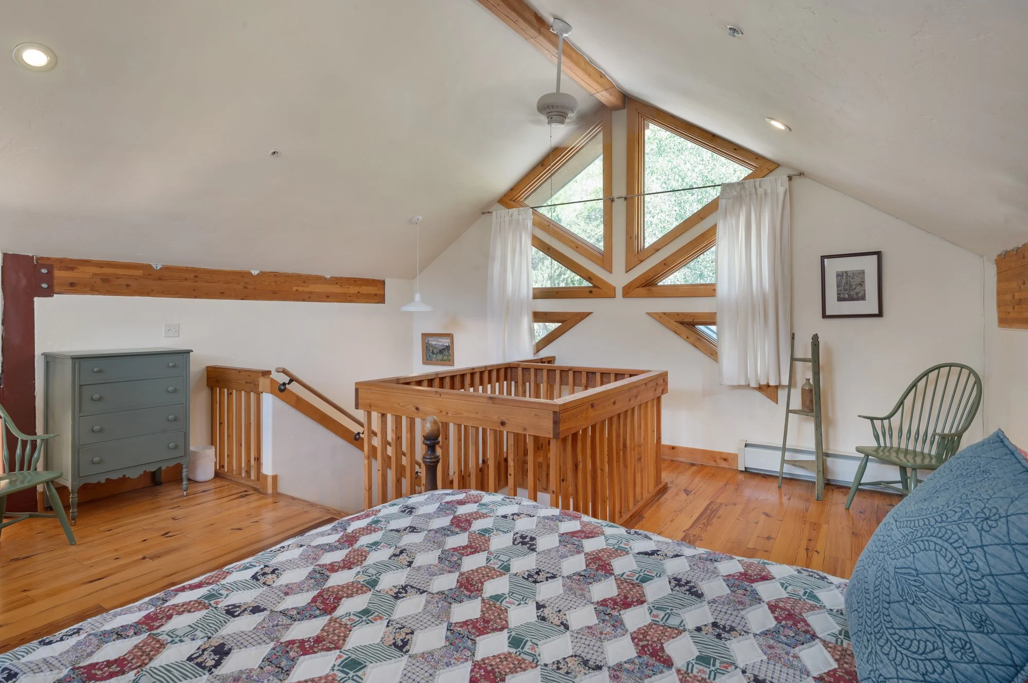 The image shows a cozy bedroom with wooden floors and a sloped ceiling. There's a bed with a colorful, patterned quilt and a blue pillow. The room features large, uniquely shaped windows with white curtains, a green dresser, two green chairs, and a s