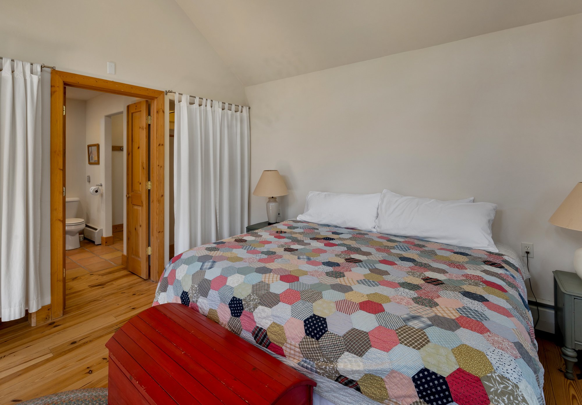 A bedroom with a large bed covered in a multicolored quilt, two pillows, wooden flooring, and two bedside lamps. There is a glimpse of a small bathroom with a toilet seen through an open doorway, with white curtains around the bathroom area.