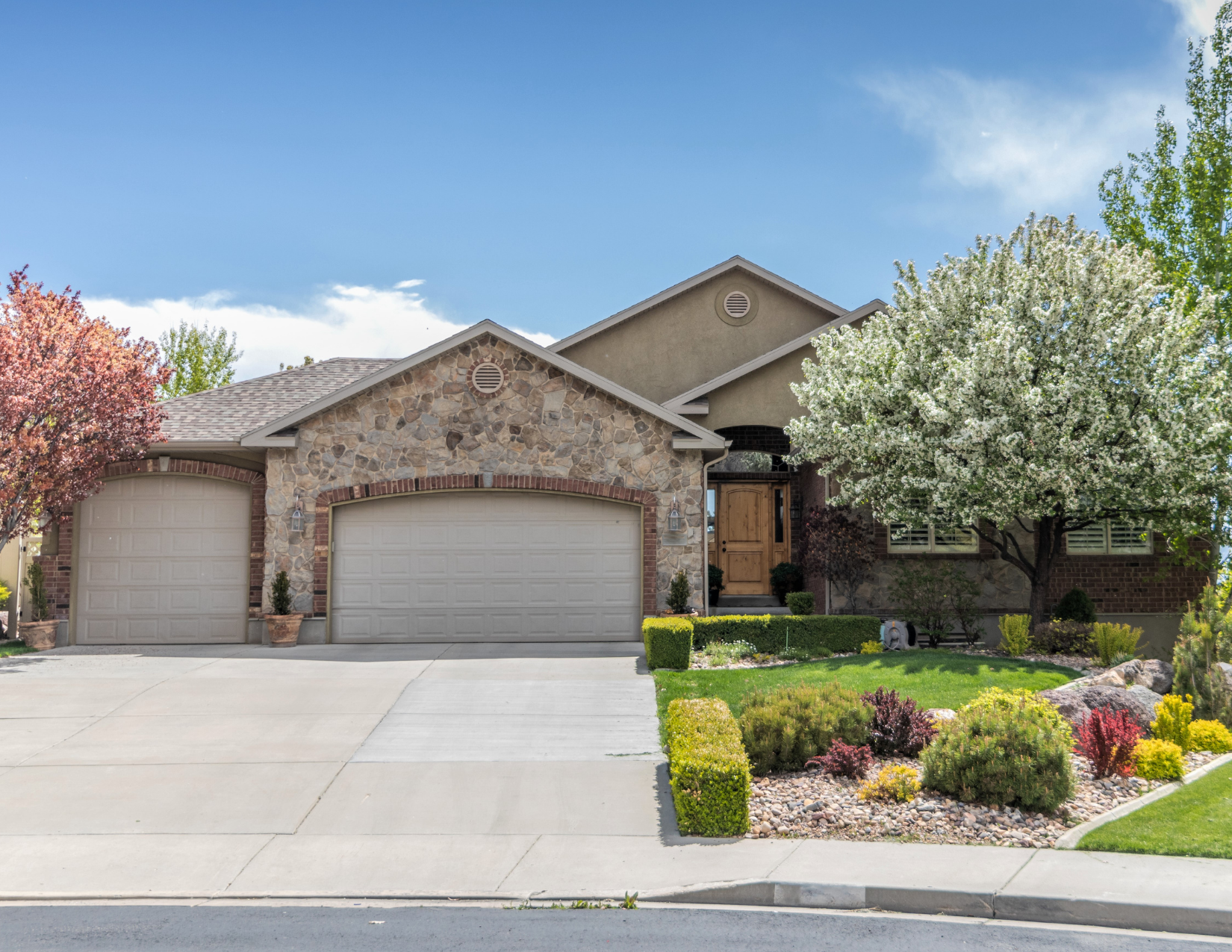 Front view of a house with stone and brick exterior, three garage doors, a wooden front door, and landscaped yard with blooming trees and bushes under a partly cloudy sky.