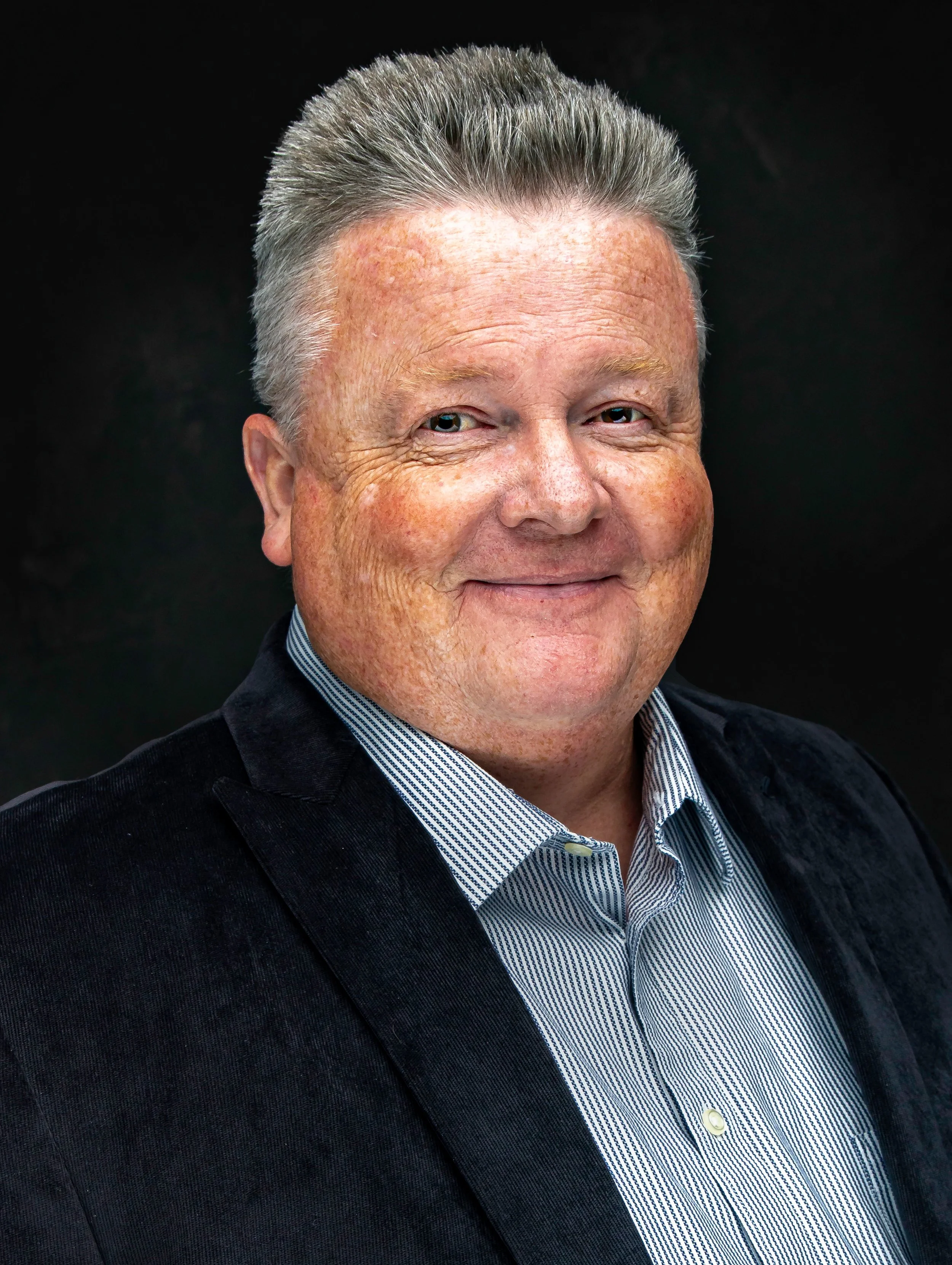 A middle-aged man with gray hair styled upward, wearing a navy blue blazer and a blue and white striped collared shirt, smiling against a dark background.