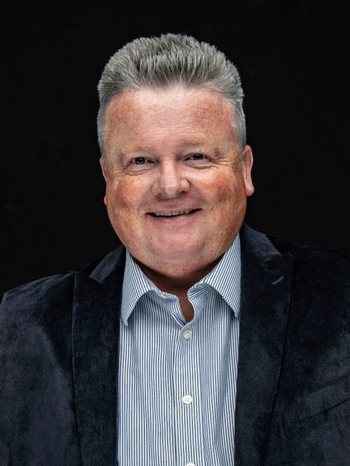 A smiling middle-aged man with gray hair in a business casual outfit against a black background.