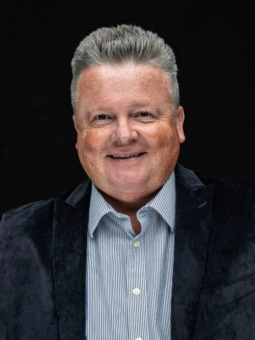 A smiling middle-aged man with short gray hair, wearing a blue striped shirt and a dark blazer, against a black background.