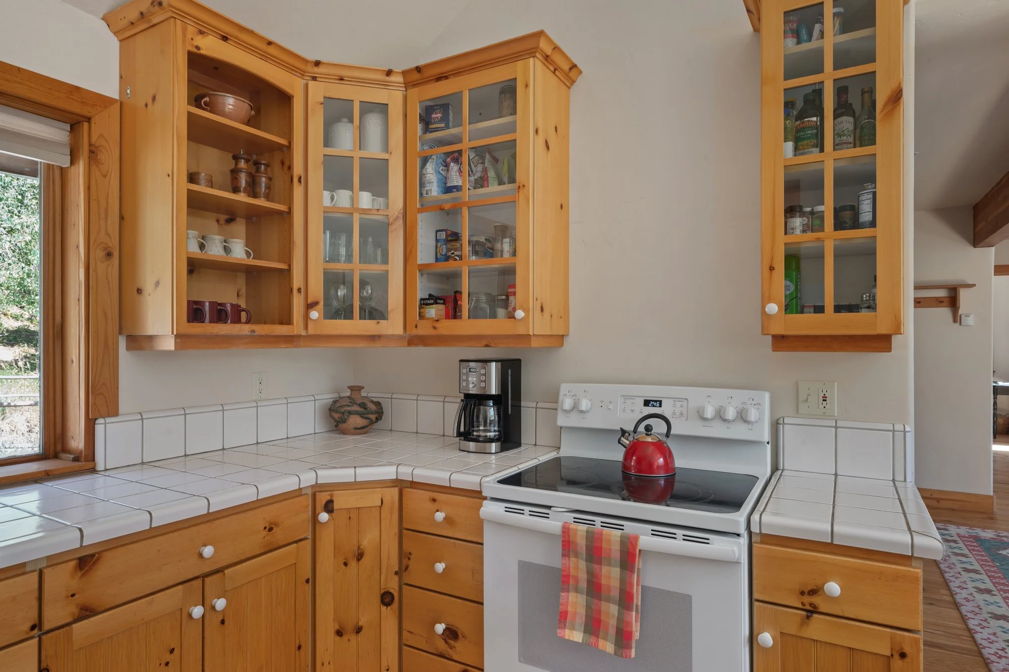 A kitchen with wooden cabinets, white tiled countertops, a window on the left, a coffee maker, a red tea kettle on the stove, and a towel hanging from the oven door.