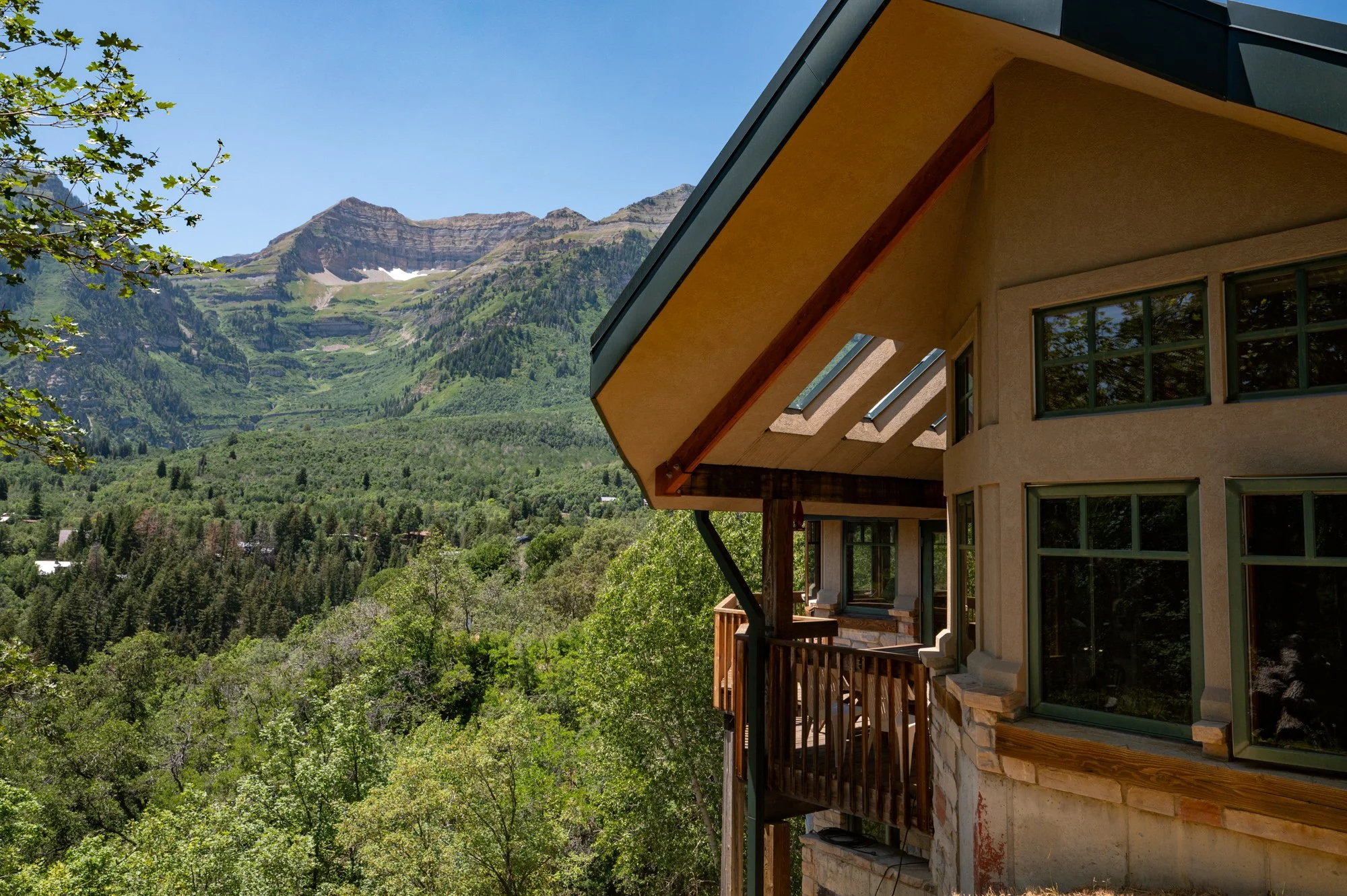 Part of a house with a balcony overlooking a lush green forest and mountains in the background during daytime.